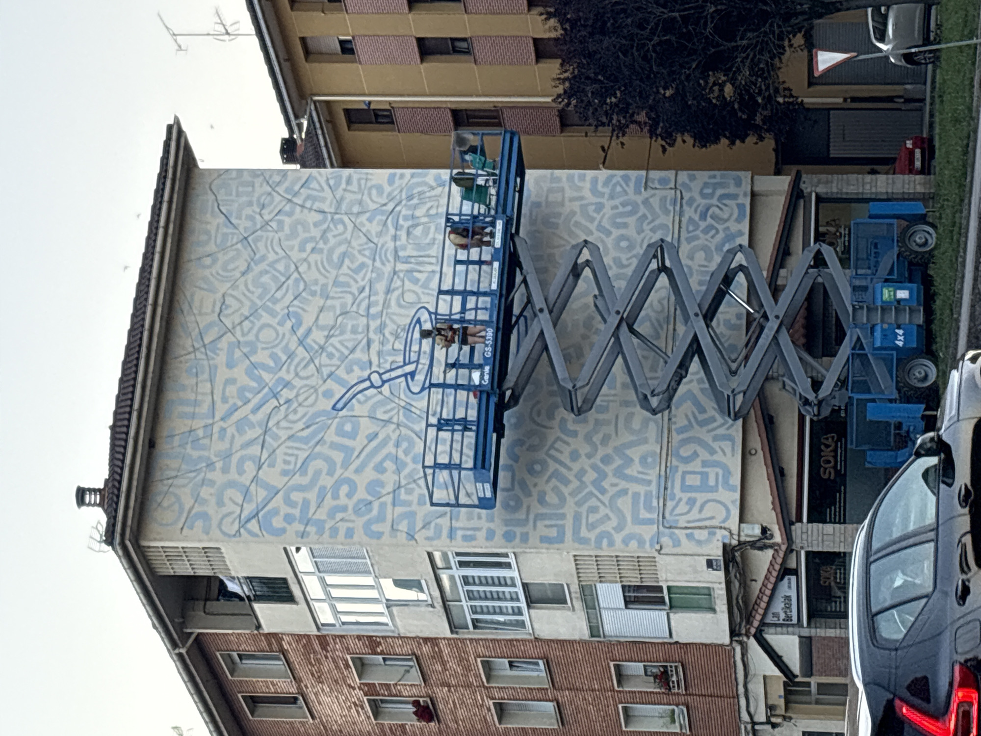 The image depicts a street scene in Mungia, Spain, featuring a building adorned with a large mural. A scissor lift is positioned in front of the mural, with three individuals standing on its platform. One individual, wearing dark clothing and a backpack, appears to be painting or working on the mural. Another individual, dressed in a light-colored shirt and shorts, is also on the platform. A third person, wearing a green shirt, is seated on a chair on the platform. The mural itself is abstract, with light blue shapes and lines covering the side of the building. The surrounding environment includes other buildings, a street with a parked car in the foreground, and a tree. The sky is overcast.