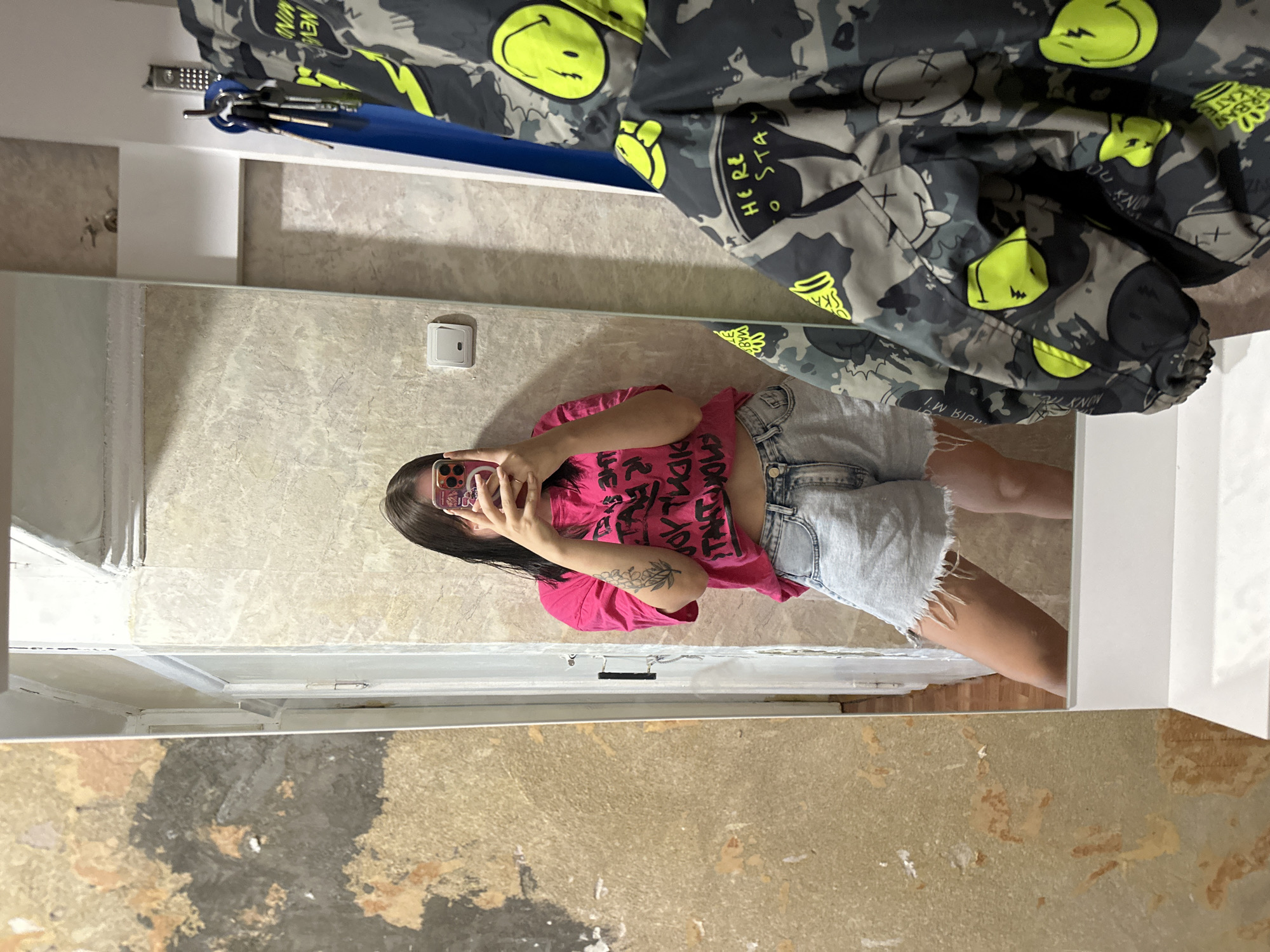 A young woman stands in front of a mirror, taking a selfie. She is wearing a bright pink cropped t-shirt with black lettering and distressed denim shorts. She has dark hair and a tattoo on her left arm. The mirror reflects a room with textured walls and a light switch. A jacket with a camouflage pattern and yellow smiley face designs hangs to the right of the mirror. The background appears to be an interior space, possibly a hallway or a dressing area, with peeling wallpaper on the left side and a doorway visible behind the woman. The scene is consistent with an apartment or residential building in the city of Chaikovsky, Russia.
