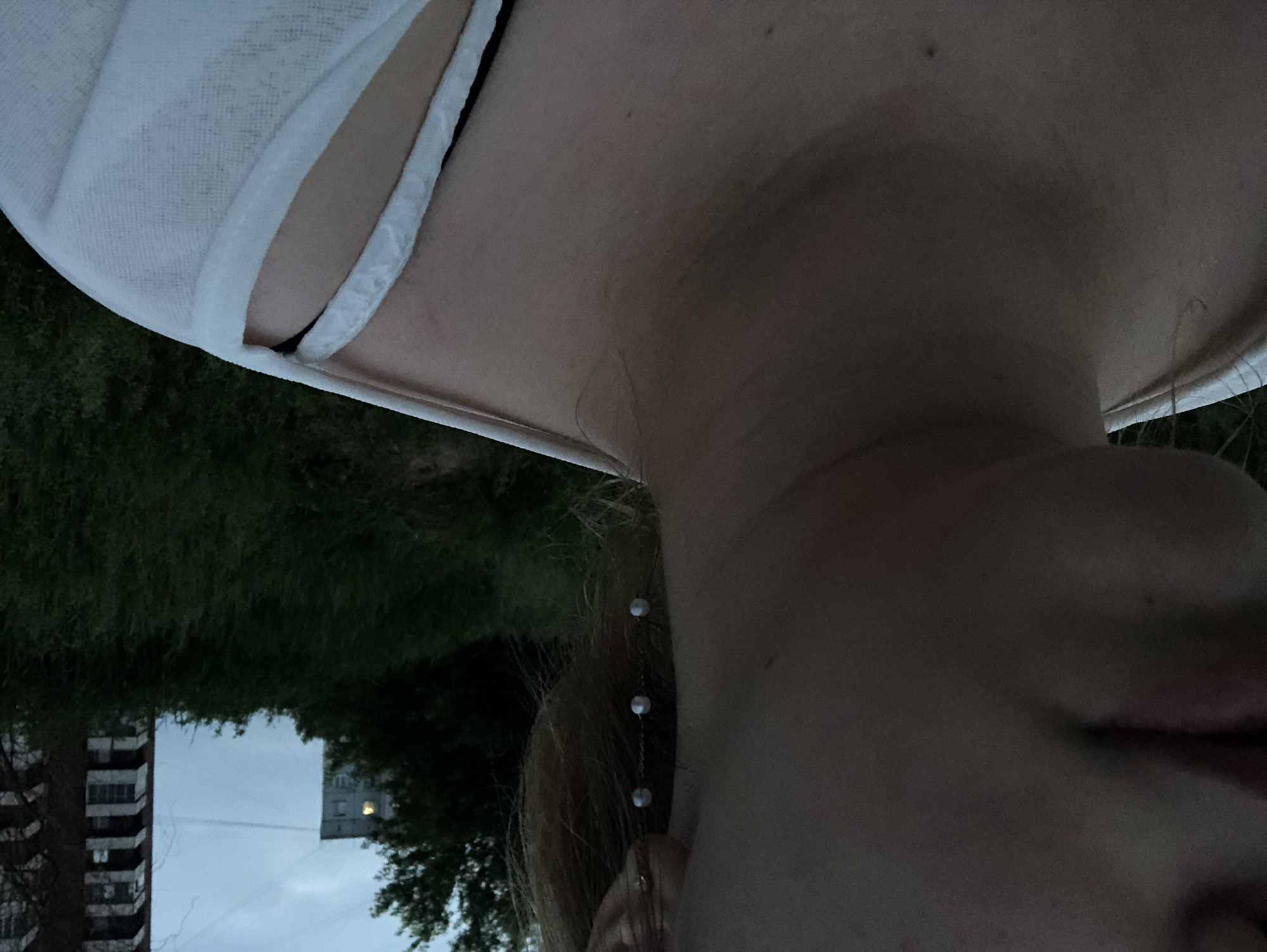 The image shows a close-up of a person's neck, shoulder, and part of their face. The person is wearing a white garment over a black strap. A delicate earring with three small pearls hangs from their ear. In the background, out-of-focus, are lush green foliage, a tree, and two apartment buildings under a twilight sky. The scene is outdoors in an urban environment.