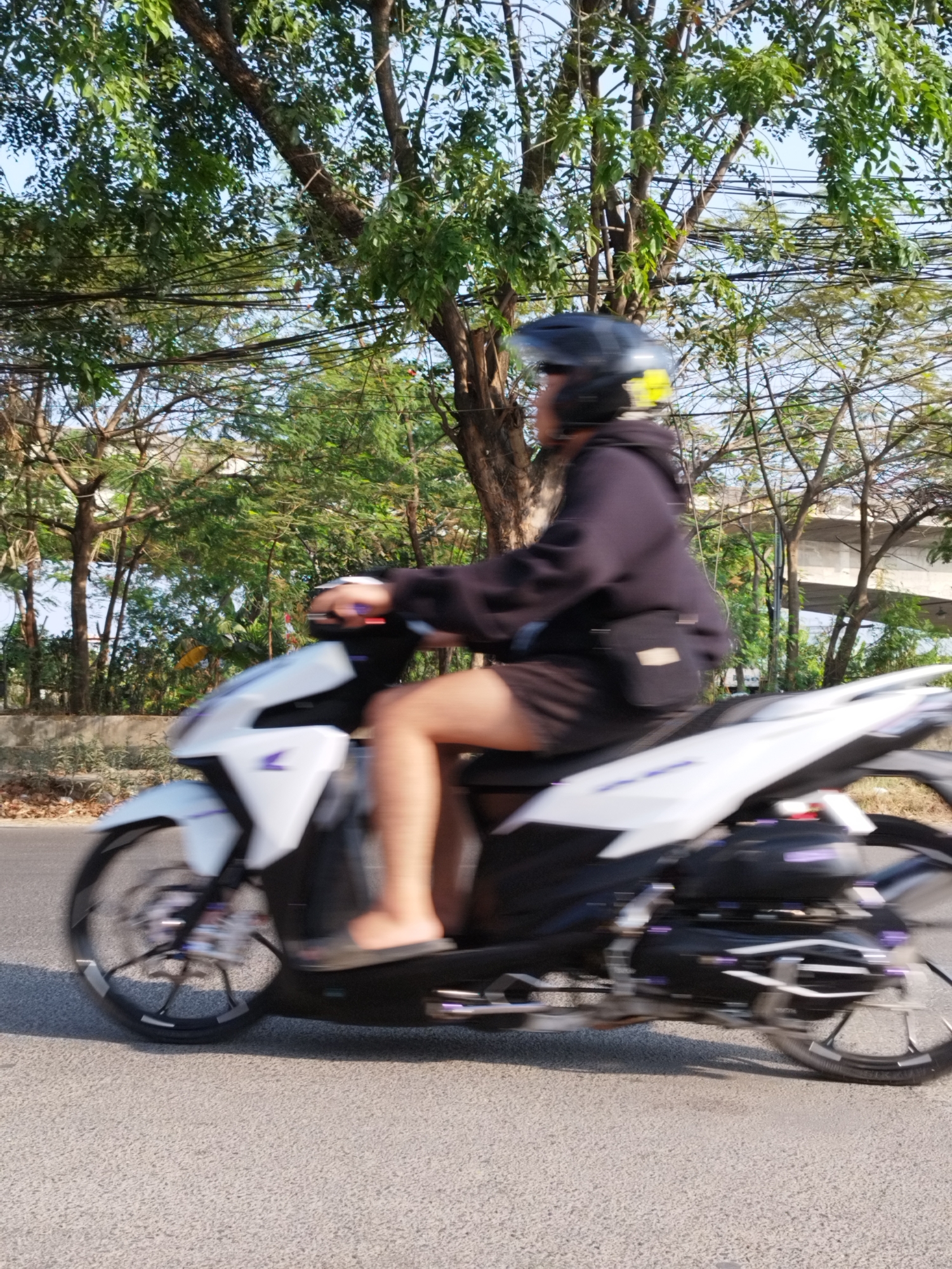 A person wearing a black hoodie, black shorts, and a black helmet rides a white scooter on an asphalt road. The person is positioned on the right side of the frame, facing left. The scooter's front wheel and body are blurred due to motion. The background features lush green trees, some with visible branches and leaves, under a clear blue sky. Overhead, power lines and a section of an elevated structure are present. The lighting suggests daytime, with sunlight casting shadows. The overall scene depicts a person riding a scooter in an outdoor, possibly urban or suburban, environment with abundant vegetation.