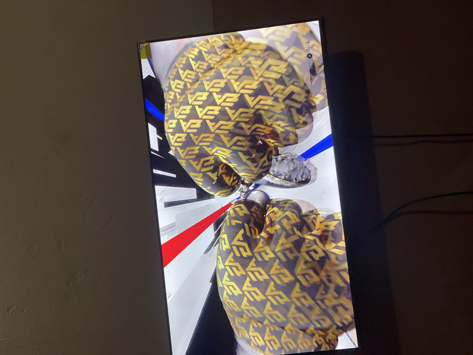 A screen displays an image of an object, likely a trophy or award, with a pattern of repeating yellow and black "EA" logos. The object is positioned amidst red, blue, and white geometric lines. "Press X to Skip" is visible in the screen's corner. The screen is angled, and the background shows a light-colored wall on the left and a dark wall with visible cords on the right.