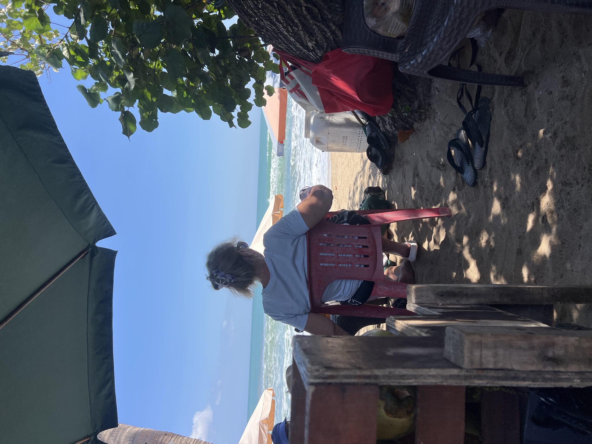 A woman with blonde hair tied back with a purple flower accessory sits on a red plastic chair facing the ocean. She is wearing a light blue t-shirt and black shorts. The chair is on a sandy beach, with the ocean and a clear blue sky in the background. Umbrellas provide shade from the sun, and a large green umbrella is visible in the foreground. To the left of the woman, a wooden structure holds several coconuts. Near her feet are a pair of black flip-flops. Behind her, a large red tote bag and a white plastic container are placed on the sand. The scene suggests a relaxed day at the beach.