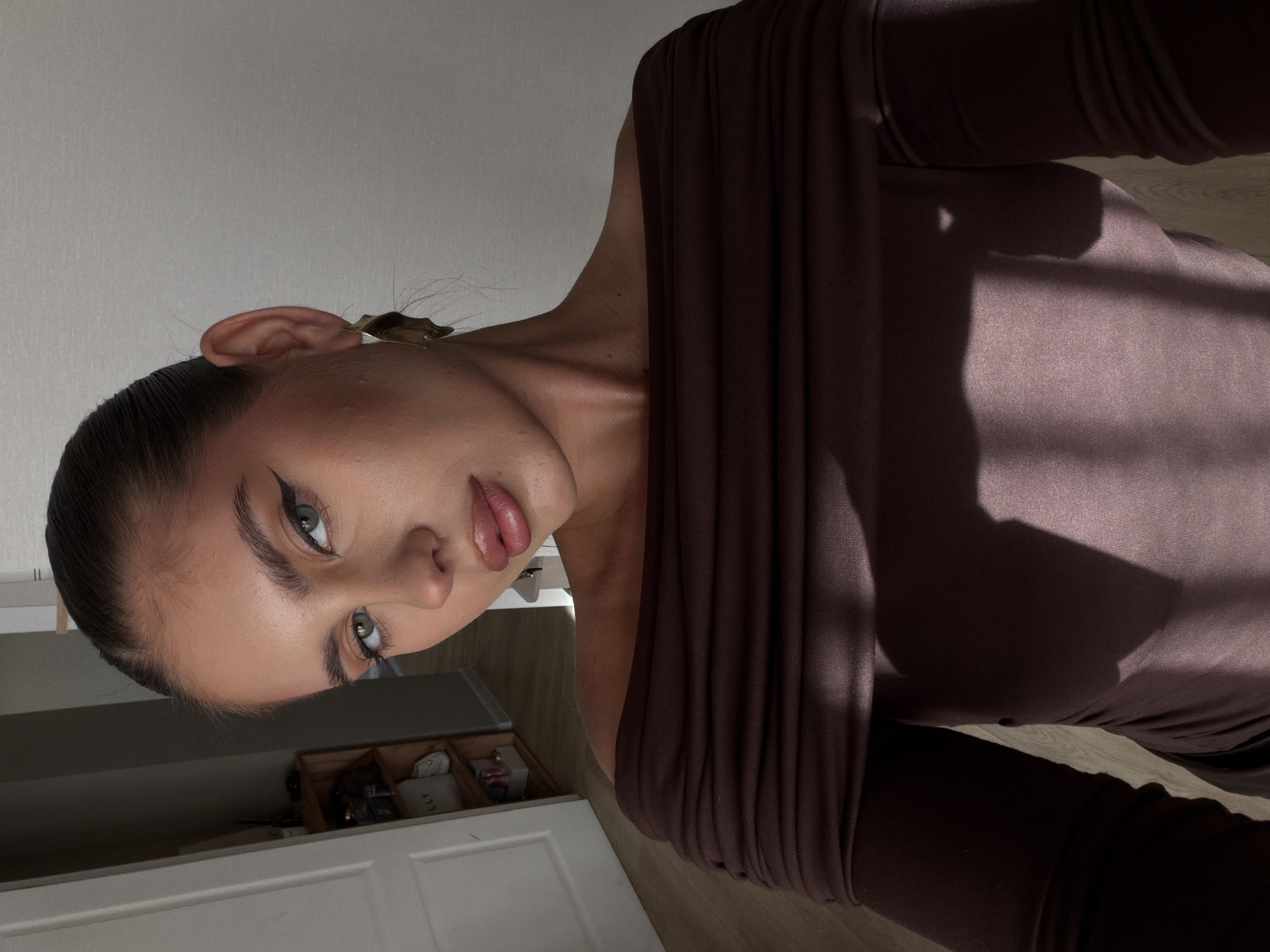 A woman with fair skin, dark hair pulled back, and winged eyeliner is the central subject of the image. She wears a brown, off-the-shoulder garment. A gold-colored hair clip adorns her hair near her ear. The background includes a white cabinet or drawer unit, and a glimpse of wooden flooring.
The image was not taken in Krasnoyarsk, Russia. There is no evidence in the photo that would place this photograph in Krasnoyarsk.