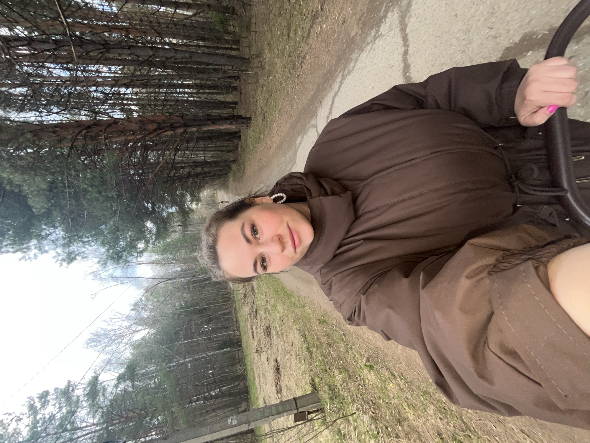 A young woman in a brown puffer jacket is outdoors on a dirt road or path lined with trees. The location appears to be in a wooded area with tall trees and sparse undergrowth. The ground cover is a mix of grass and dried leaves. She is holding a black, circular object, possibly part of a bicycle or stroller. Her fingernails are painted pink. The location is Chaikovsky, Russia.