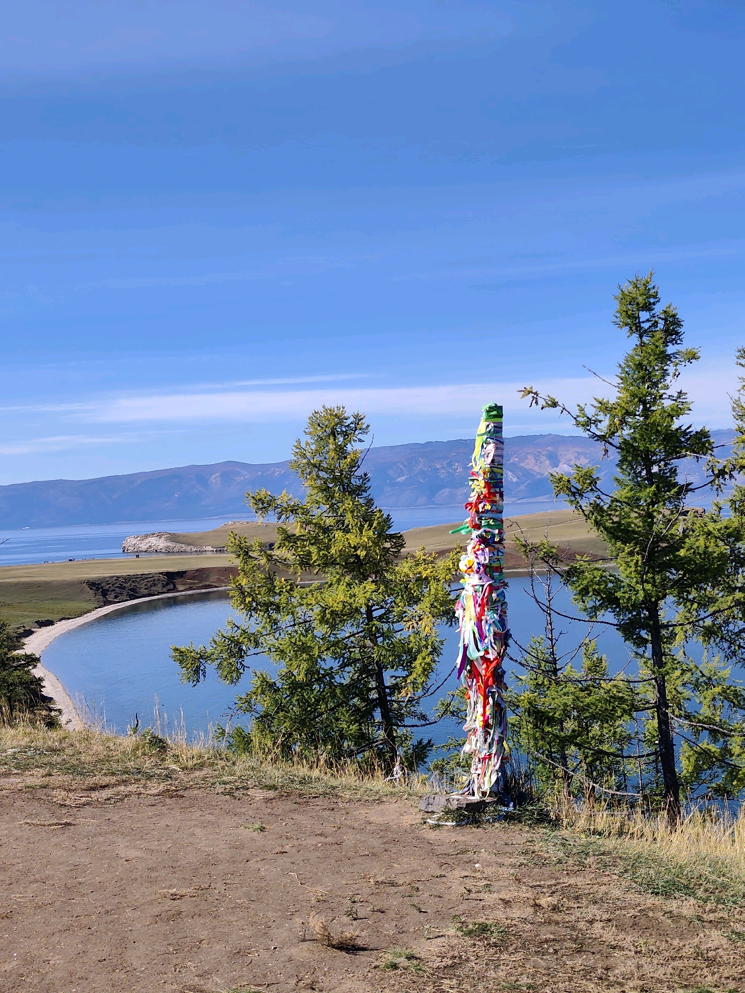 A totem pole adorned with colorful ribbons stands on a grassy knoll overlooking a calm, blue body of water. The totem pole is a central focus, its vibrant ribbons contrasting with the natural scenery. Behind it, a bay curves inward, with a sandy beach along its edge. The water stretches out to meet distant, hazy mountains under a clear, blue sky. Two evergreen trees frame the right side of the image, their branches reaching towards the sky. The foreground consists of a dirt path with dry grass. The scene appears to be the village of Kharantsy, Russia, a location known for its proximity to Lake Baikal and its spiritual sites.