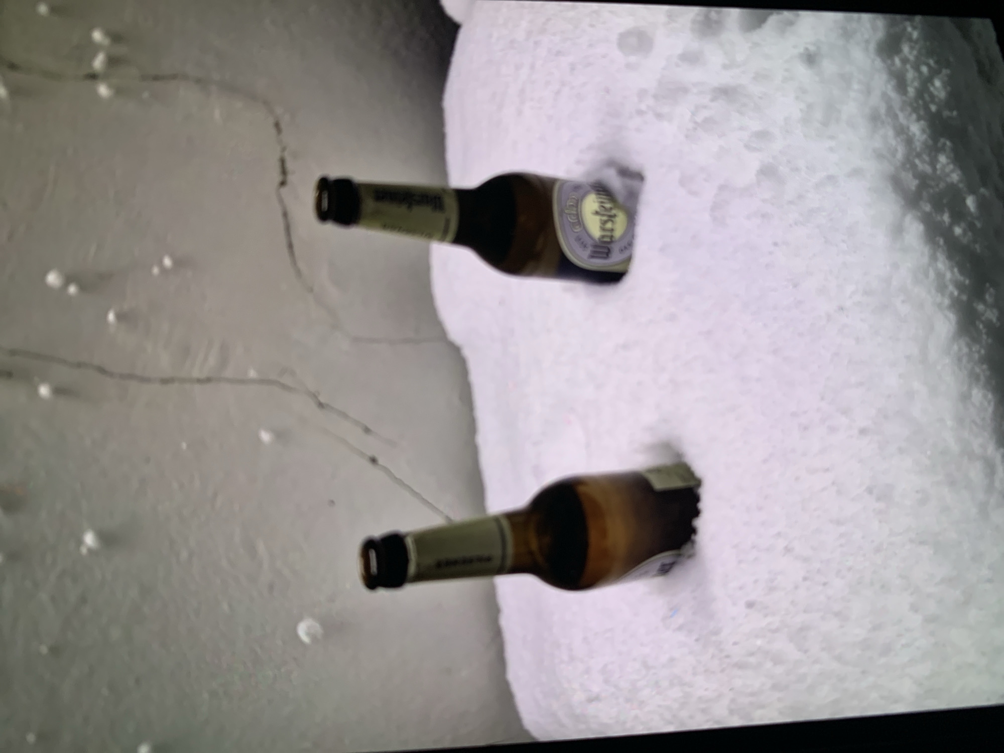 The image displays two beer bottles horizontally embedded in a thick layer of snow or ice. The bottles appear to be chilling in the cold environment. The snow or ice takes up the right and bottom portions of the frame, with a lighter, possibly frozen or snowy surface extending to the left. Small, scattered white dots and faint lines are visible on this lighter surface, possibly indicating ice formations or texture.
On the upper bottle, the label "Warsteiner" is clearly visible, accompanied by a crown logo. The neck label also appears to have text, likely "Warsteiner" as well. The bottle itself is dark brown glass with a green neck label. The lower bottle is similarly dark brown with a green neck label, but the main label's text is less distinct, though it seems to match the Warsteiner branding of the upper bottle. The scene suggests an outdoor winter setting where beverages are being kept cold naturally. No people are visible. The lighting is bright, possibly indicating daytime.
