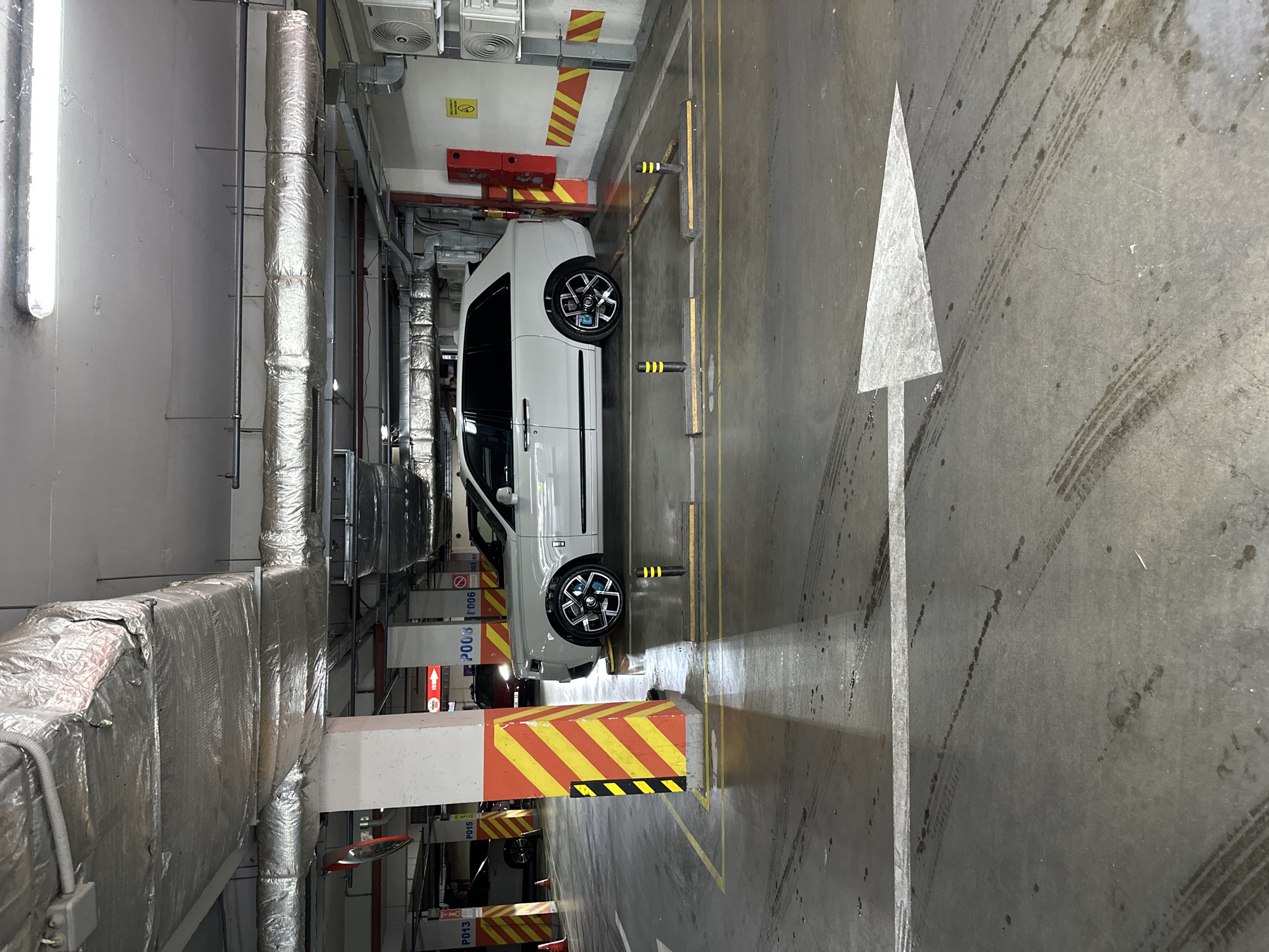 A white Rolls-Royce Cullinan SUV is parked in a covered, multi-level parking garage in Chișinău, Moldova. The vehicle is positioned between concrete pillars painted with diagonal yellow and red stripes. The floor of the garage is concrete, showing tire marks and some damp spots. In the foreground, a large white arrow points to the right, indicating a traffic direction. The garage ceiling features exposed ductwork and lighting fixtures. Other cars and parking signs are visible in the background.