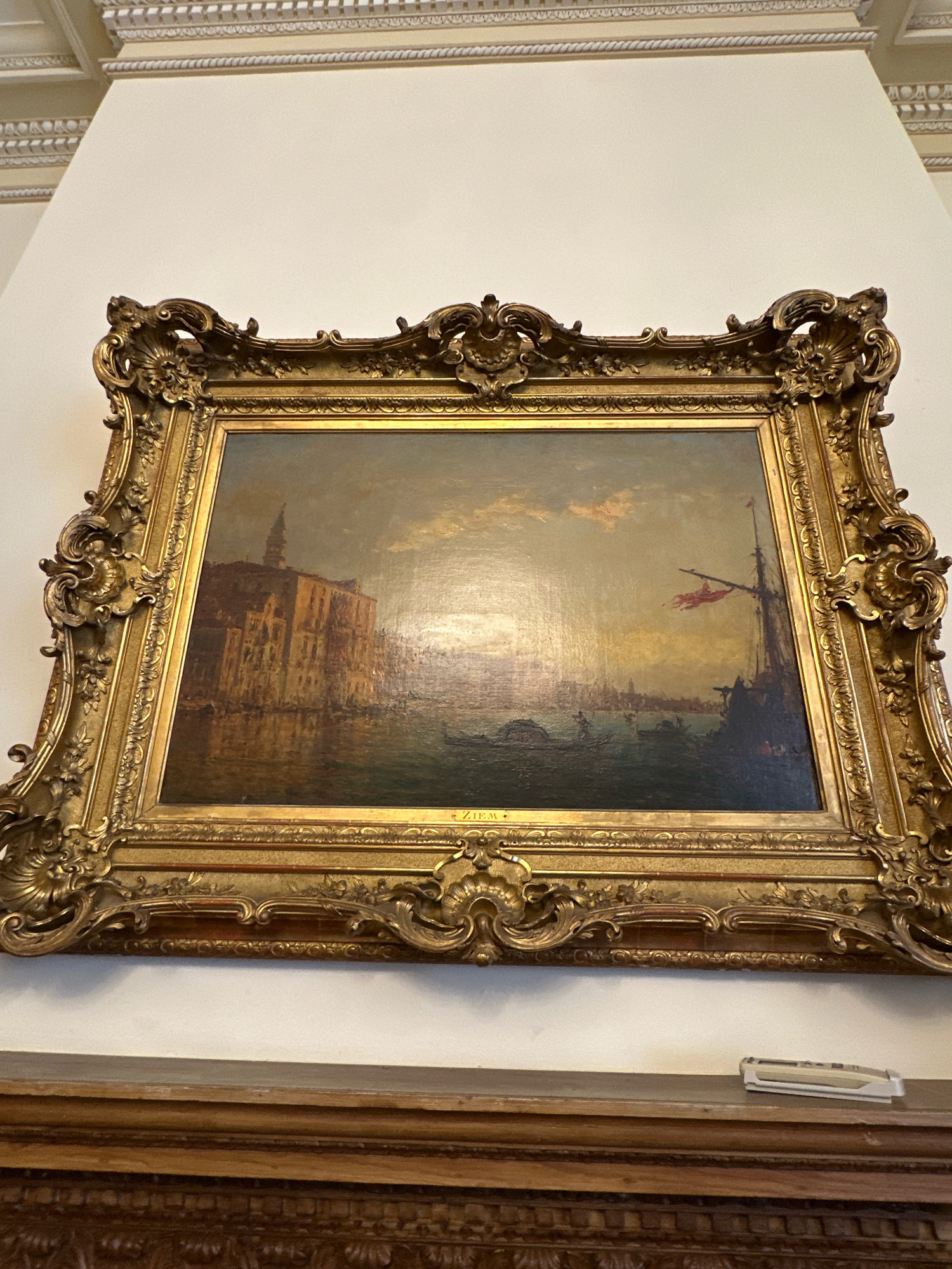 The image shows a painting in an ornate gold frame, displayed on a white wall above a wooden surface. The painting depicts a Venetian scene with buildings lining a canal, a boat, and a ship with a red flag. The sky is hazy with sunlight reflecting on the water. The artwork is signed "ZIEM" in the lower right corner. The wall features decorative molding at the top. The wooden surface below the painting has carved details. A small white object, possibly a label or a card reader, is placed on the wooden surface. There is no visible indication of this location being in Belgrade, Serbia.