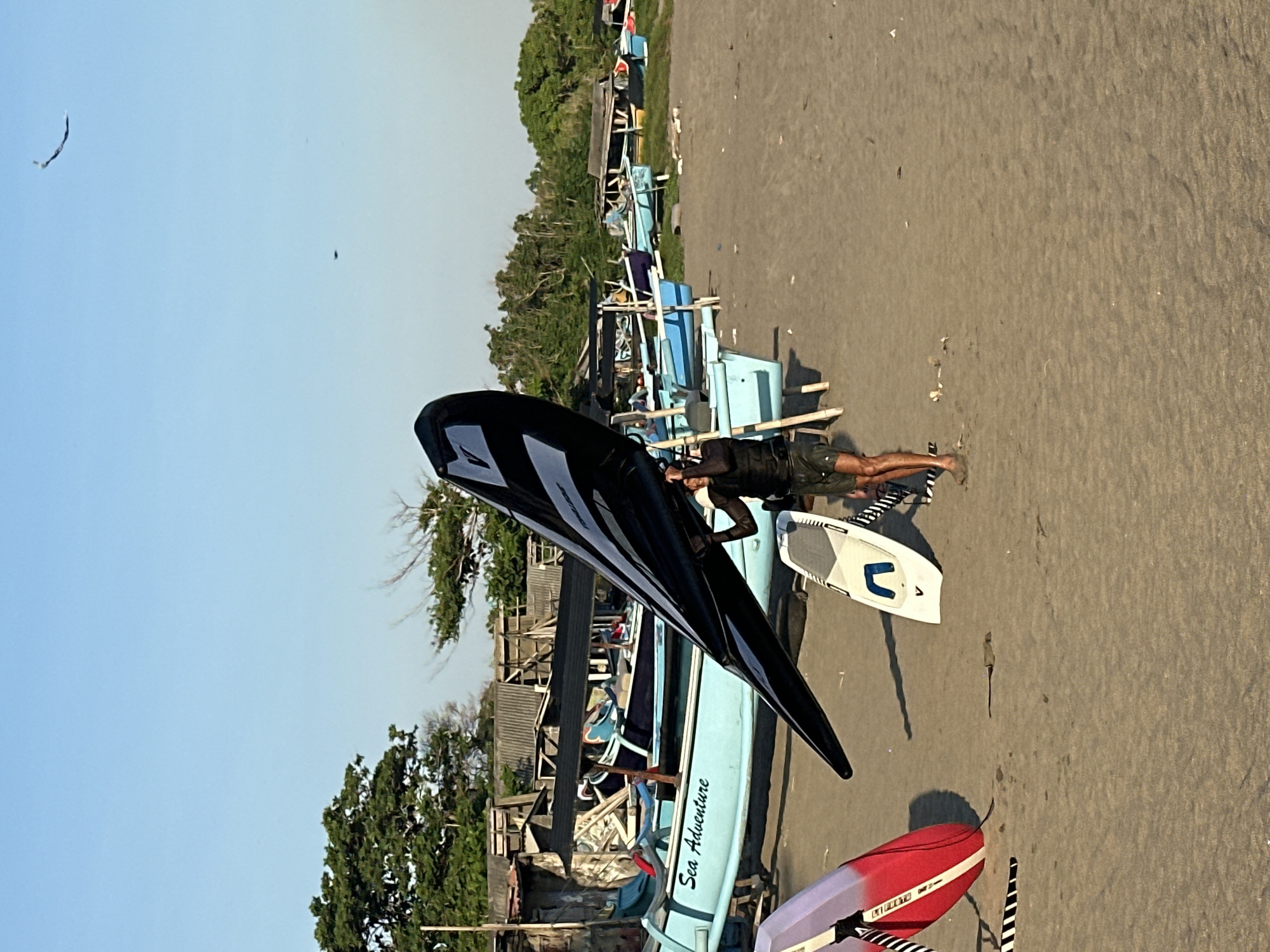 A person is lying on a sandy beach in North Kuta, Indonesia, near several boats and water sports equipment. The person is wearing dark shorts and is partially covered by a large, black and white wing, likely for wing foiling. To the right of the person, a white board with blue accents is visible, and further down, a red and white board rests on the sand.
In the background, a line of light blue boats is pulled up onto the beach. Some of these boats have structures built over them, possibly for shade or storage. Behind the boats, there are trees and simple buildings, suggesting a coastal village or a watersports rental area. The scene is bathed in bright sunlight, indicating it is daytime, likely late afternoon or early morning due to the long shadows. The sky is clear and blue. The overall atmosphere appears relaxed and peaceful, with the equipment suggesting recreational activities.
The text "Sea Adventure" is visible in white lettering on one of the light blue boats.
