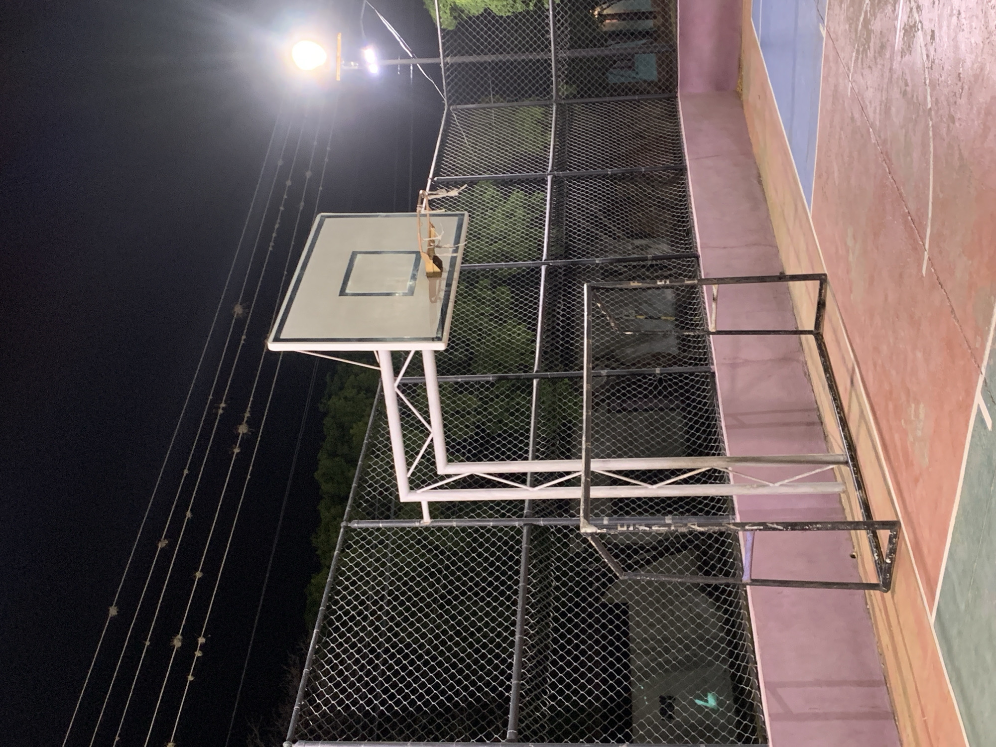 The image captures an outdoor basketball court at night in Santa Ana, Venezuela, illuminated by bright artificial lights. The primary subject is a basketball hoop featuring a white backboard and a metal rim, which notably lacks a net. The hoop assembly is supported by white metal framework. The court surface itself is distinctive, showing signs of wear and tear, and is painted in multiple colors including sections of red-orange, blue, and green, with white lines marking boundaries.
The court is enclosed by a tall chain-link fence, visible running across the middle and right side of the frame, suggesting a dedicated sports facility. Beyond the fence, a dark background with hints of trees or foliage confirms the nighttime setting. In the upper left, several utility wires or cables stretch across the dark sky, with small, irregular clumps of material visible clinging to them. Adjacent to the court on the right, there is a low barrier or wall painted a light pinkish-purple, possibly demarcating the court from a walkway or other ground. The bright overhead lights create strong glows and reflections, indicating the sole source of illumination. There are no people visible, and no activity is taking place on the empty court. No text is visible in the image.