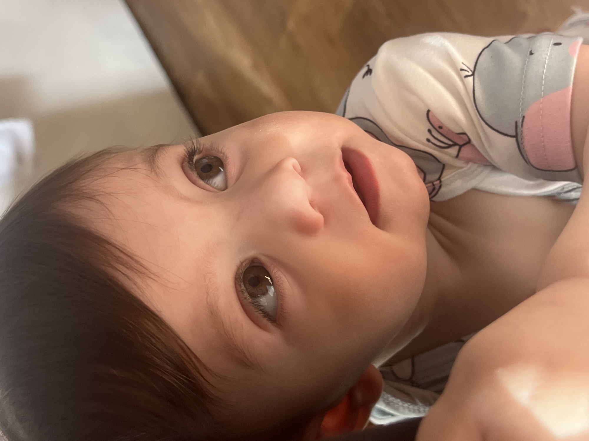 A close-up, upward-angled shot shows a young child with brown hair and eyes, wearing a patterned white and gray shirt with pink accents. The child's face is the primary focus, with a slightly open mouth. Parts of the child's upper body and arm are visible in the lower right of the frame. The background is blurred and out of focus, showing a light tan color at the top left. There is no indication of Elena, Bulgaria in the image.