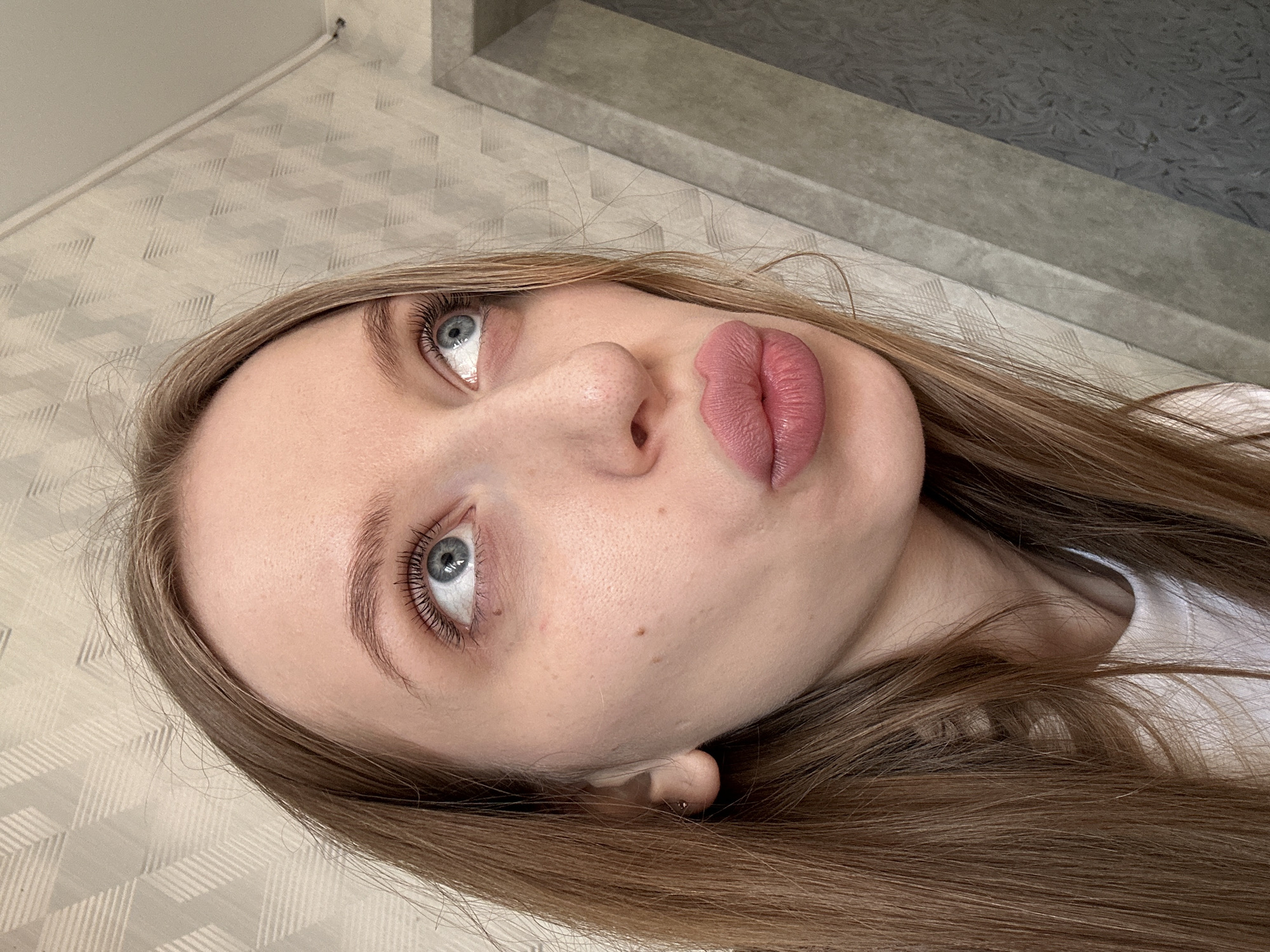 A close-up, angled portrait of a fair-skinned woman with long, light brown hair. She is looking upwards, with light blue eyes visible. Her lips are pursed in a kissing gesture, and they are a light pink color. She has a mole near her lower right eye. Her hair is straight and smooth, falling around her face. The background consists of textured wallpaper and a gray, possibly concrete, ledge. No information suggests this image was taken in Chaikovsky, Russia.