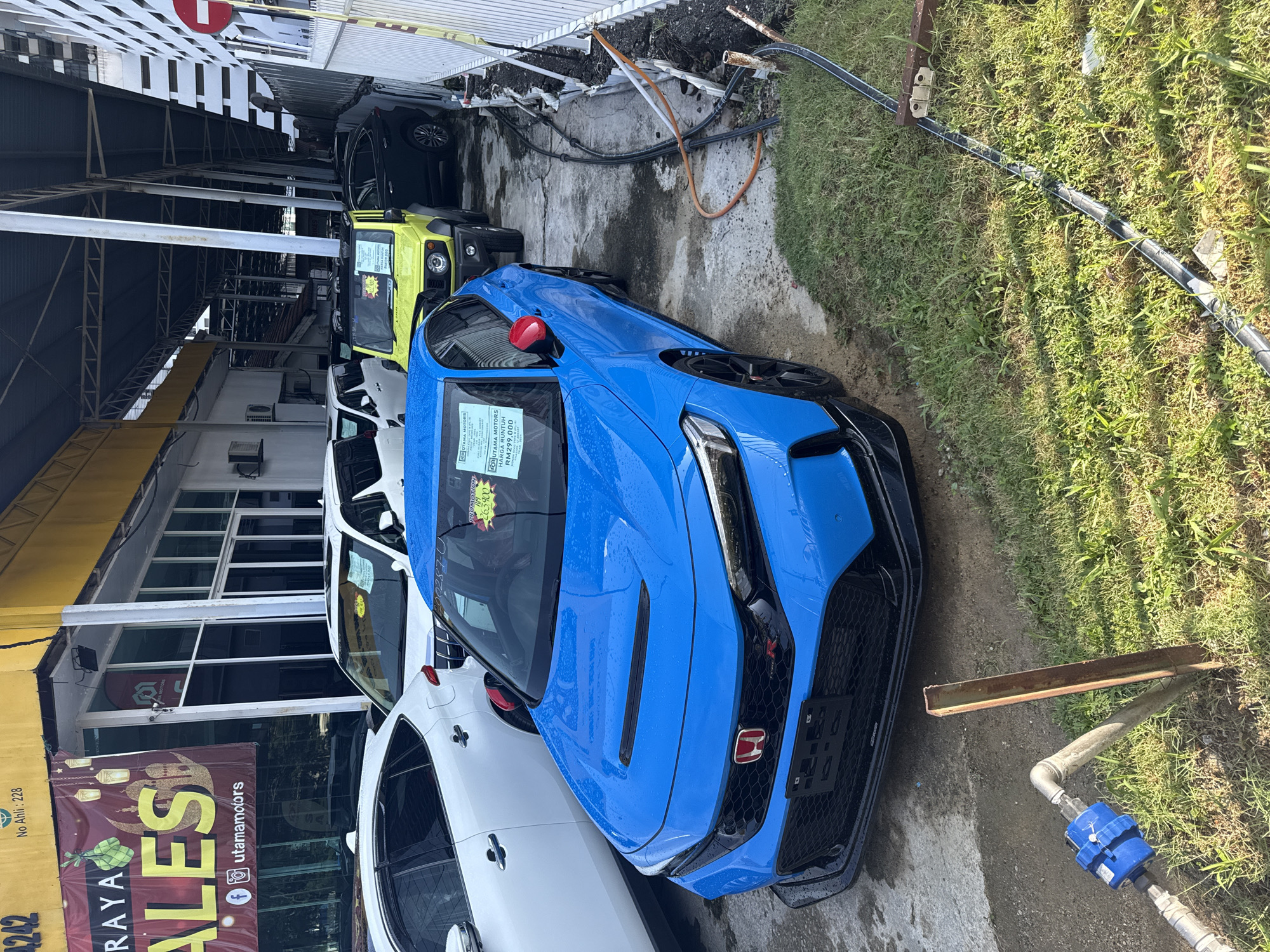 The image shows a blue Honda Civic Type R parked at a car dealership named Utama Motors. The dealership is located in Kuala Lumpur, Malaysia. The car is on display with a sales price tag on the windshield. The price is 299,000 Malaysian Ringgits, which is a discount from 263,900 Malaysian Ringgits. Other vehicles are visible in the background. A metal pipe with a blue valve runs along the ground near the front of the car. The dealership is located at No Ahli: 228
4242. A banner that reads "RAYA SALES" is visible on the dealership's building.