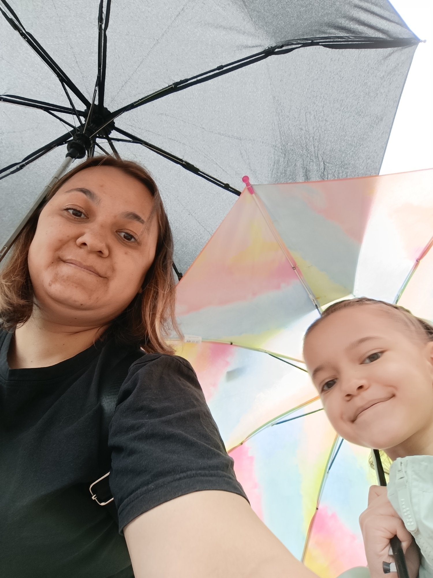 A selfie shows a woman and a young girl under umbrellas. The woman is on the left with medium-length brown hair, wearing a black t-shirt. The girl is on the right, partially hidden under a colorful umbrella. Above them, there is a larger black umbrella. The background is out of focus, suggesting an outdoor setting. The location is Chaikovsky, Russia.