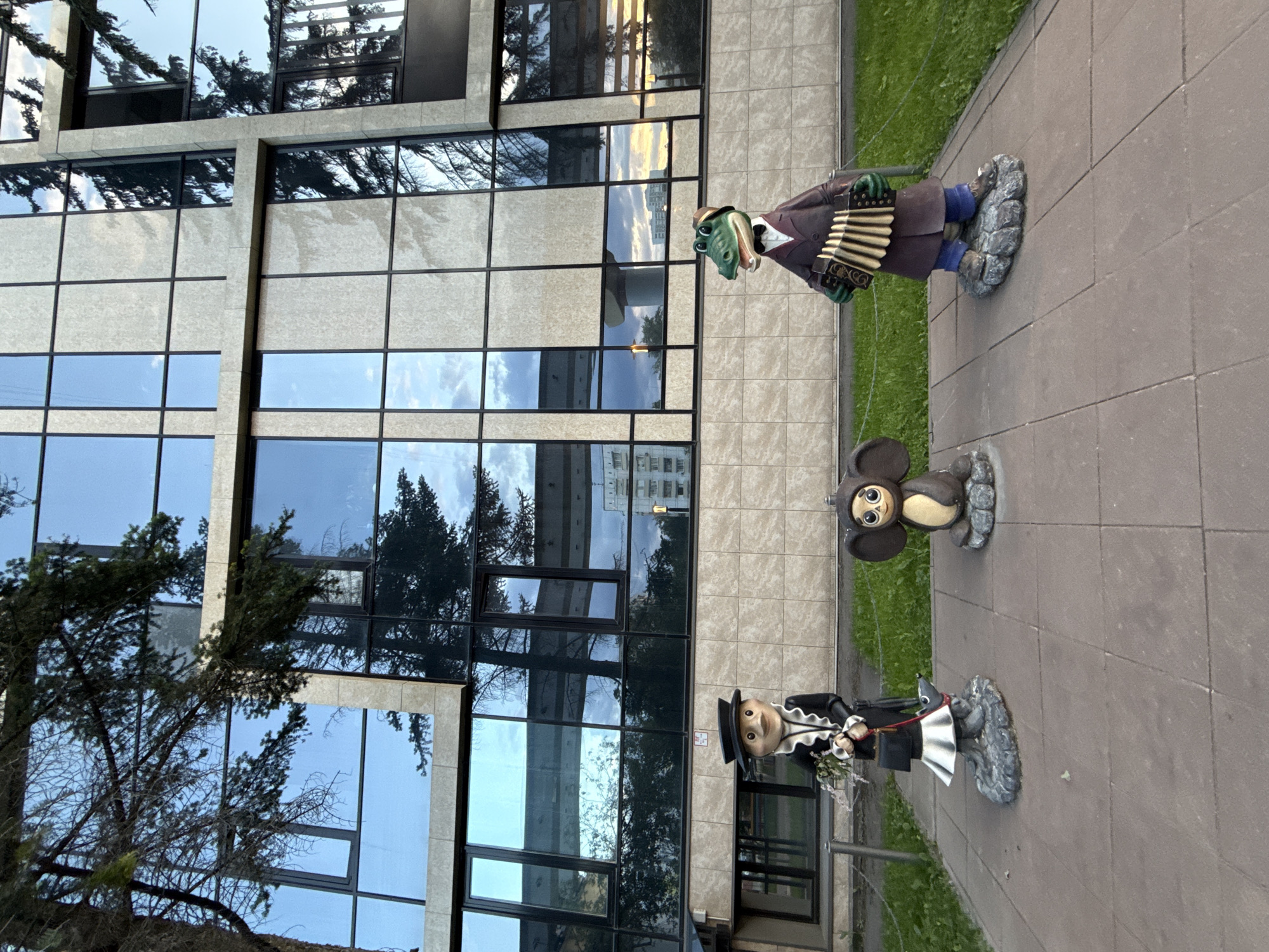 The image displays an outdoor scene in Moscow, Russia, featuring three statues of well-known characters from the Soviet/Russian animated series "Cheburashka and Friends." The statues are arranged on a light grey paved walkway, with a strip of green grass visible to their right.
From right to left, the first statue is Gena the Crocodile. He stands upright, dressed in a brown suit jacket, a white shirt with a red bowtie, and a brown fedora hat. He is actively playing a green and white accordion. His feet are stylized as a grey, textured base.
Beside Gena is Cheburashka, a small, brown, and cream-colored creature with large ears and round eyes, known for his endearing expression. He is seated on a similar grey, rock-like base, looking straight ahead.
Further to the left is the statue of Old Woman Shapoklyak. She is depicted standing, wearing a black dress with a distinctive white ruffled collar, a black top hat, and a black handbag in her right hand. By her feet, on a leash she holds, is her companion, the small rat Lariska, sitting on its own rock-like base.
In the background, a modern multi-story building with a facade composed of large, rectangular, highly reflective glass windows and light-colored stone panels dominates the left side of the frame. The windows mirror the sky, showing a mix of soft blue and white clouds, along with reflections of trees, some with sparse branches, suggesting either late autumn or early spring. The reflections also include glimpses of other distant buildings, indicating an urban environment. A large tree with dark branches is partially visible on the far left. The overall impression is of public art depicting beloved characters against a contemporary architectural backdrop. The lighting suggests it is daytime, possibly morning or late afternoon, given the soft quality of the light reflected in the windows. No discernible text is visible on the statues or the building.