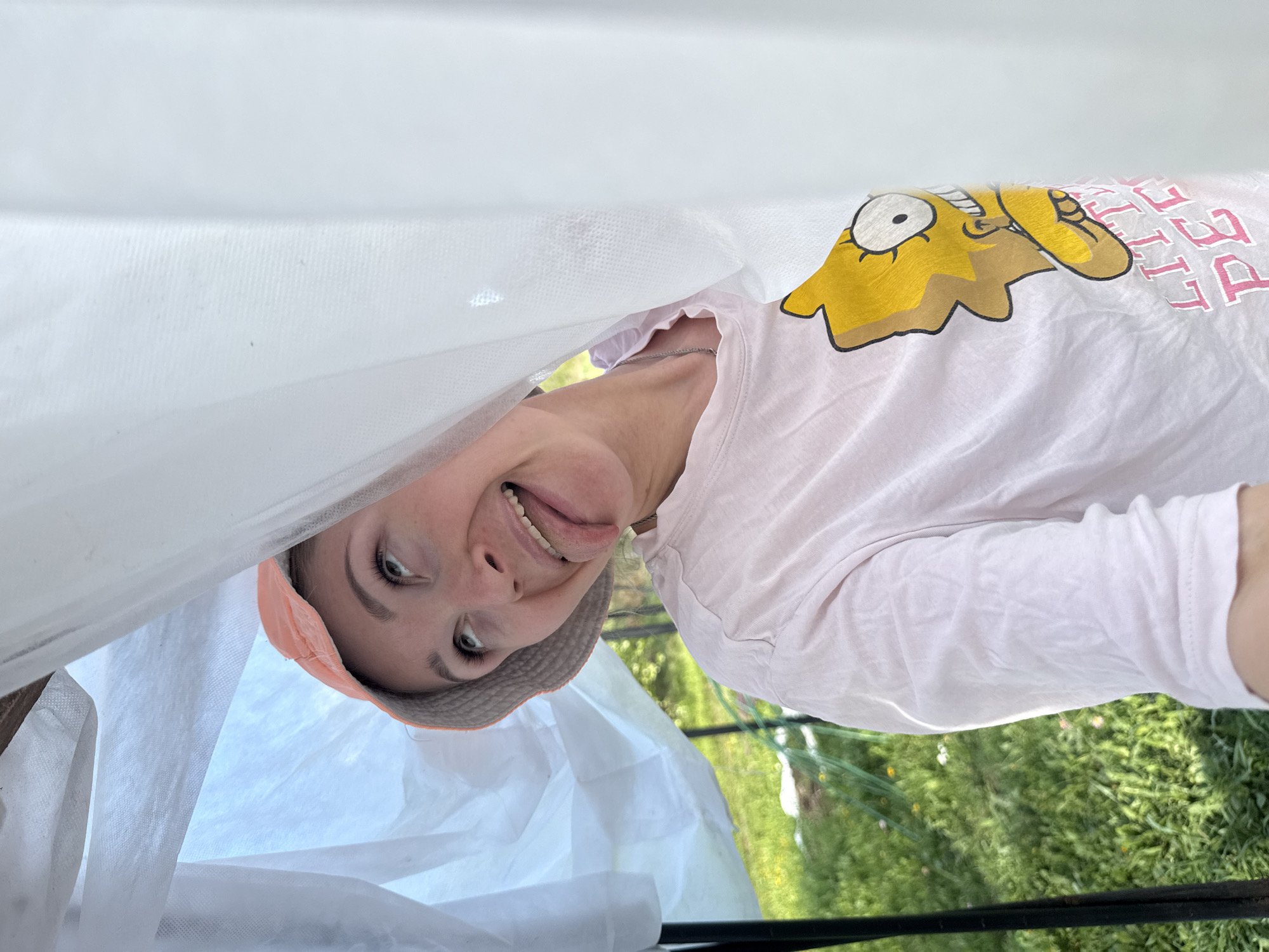 The image shows a person wearing a light pink t-shirt with a Homer Simpson graphic, standing inside a structure covered with a white fabric material. The person is wearing a hat and sticking their tongue out playfully. The background appears to be a grassy area with some vegetation, possibly a garden. There is no verifiable information about the location being in Nizhnyaya Chermoda, Russia.
