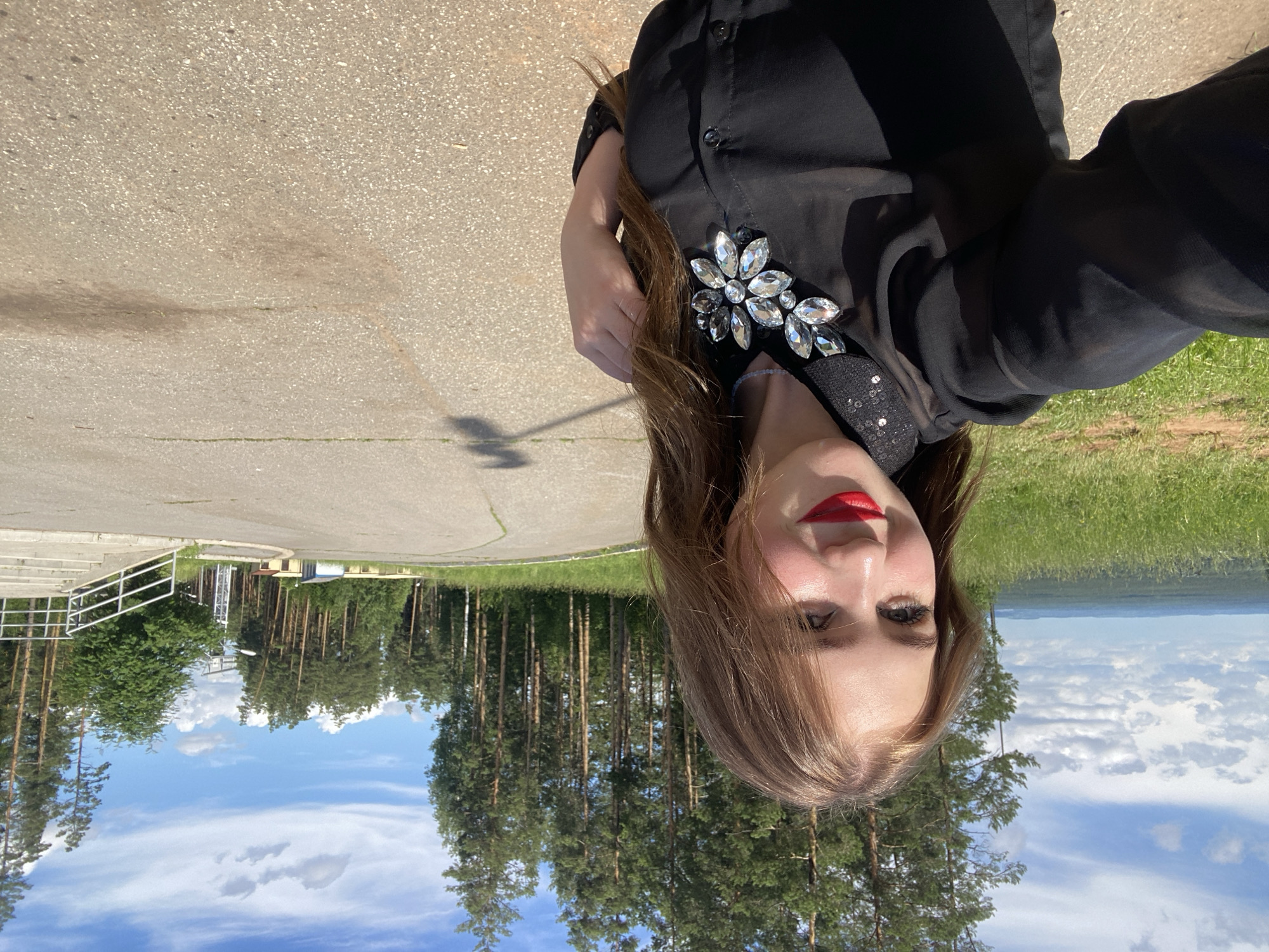 A young woman with long, light brown hair and bright red lipstick smiles at the camera, holding her left arm up to take a selfie. She is wearing a sheer black long-sleeved blouse with a sparkly collar and a large, ornate crystal brooch on her chest. The background is a paved road curving around a grassy hillside dotted with tall pine trees. In the distance, a hazy landscape of green hills and trees stretches under a bright blue sky with scattered white clouds. To the right of the road, a concrete staircase leads up to a railing, and beyond that, several small, simple buildings are visible. The scene appears to be a scenic overlook or a park area in a rural or suburban location.