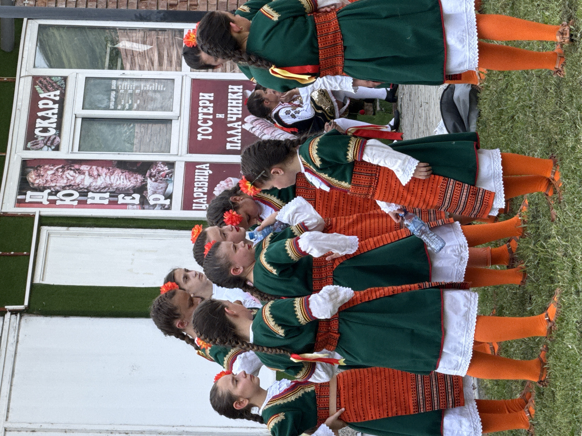 In Pordim, Bulgaria, a group of young girls dressed in traditional Bulgarian folk costumes stand in an outdoor setting. The costumes consist of green dresses with red and gold trim, paired with orange skirts, white blouses, and orange leggings. Many of the girls have their hair in braids adorned with red flowers. They are gathered in front of a building with signs in Bulgarian. One sign displays images of meat and the word "CKAPA", suggesting a food establishment. Another sign reads "ЦАРЕВИЦА TOCTEPH И ПАЛАЧИНКИ". The ground is covered in green grass. The overall atmosphere appears to be a cultural event or performance.