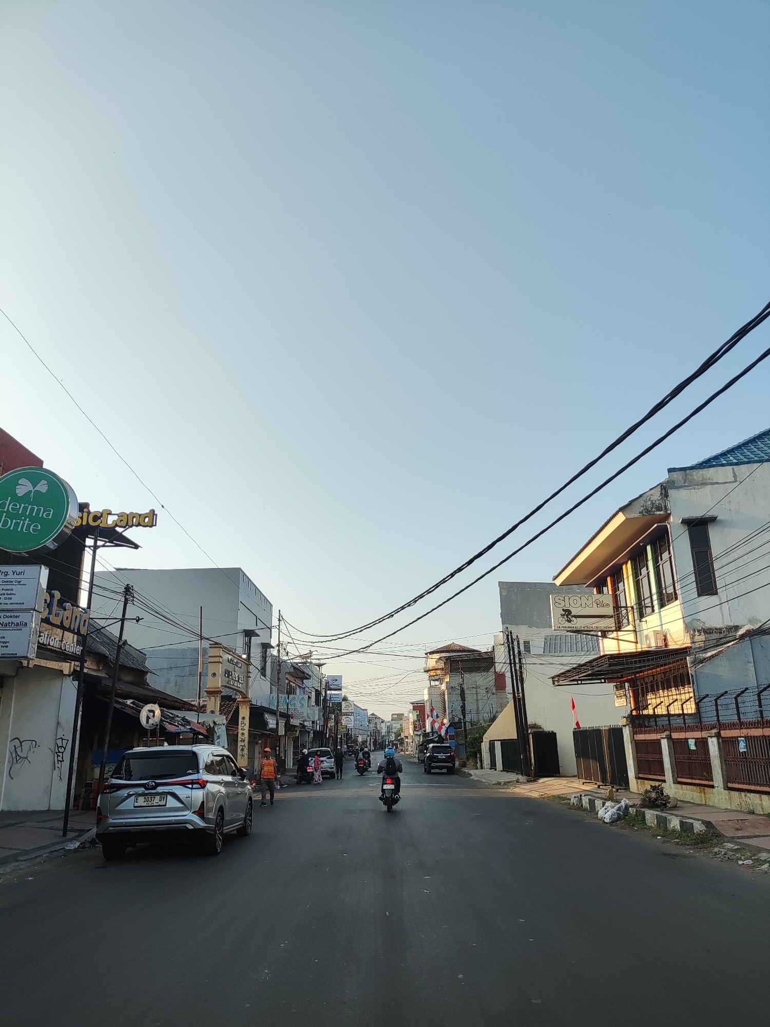 The image shows a street scene in Cirebon, Indonesia. The street is lined with buildings on both sides, with a clear sky above. Several vehicles are visible, including a silver SUV parked on the left, a motorcycle in the distance, and another motorcycle on the right side of the road. People can be seen walking and riding motorcycles. Power lines are strung across the street, obscuring parts of the sky and buildings. Signs for businesses such as "derma brite" and "SION Bike" are visible on the buildings. The overall impression is a typical urban street during daylight hours, possibly late afternoon due to the soft light.