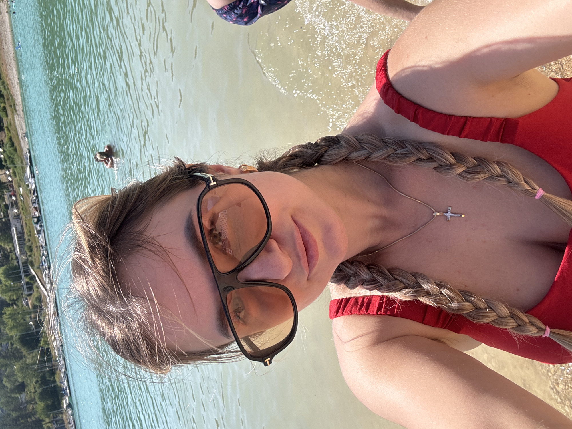 A woman with blonde hair in two braids, wearing oversized sunglasses and a red swimsuit, is taking a selfie at the edge of a body of water. The water is clear and calm, with a sandy or pebbly bottom visible. In the background, there is a shoreline with trees and buildings, and several boats are docked or on the water. Another person is visible in the water in the distance, swimming. The sky is blue and the overall atmosphere appears to be a sunny day at a lake or reservoir, possibly a recreational area. The location is identified as Maisod, France.