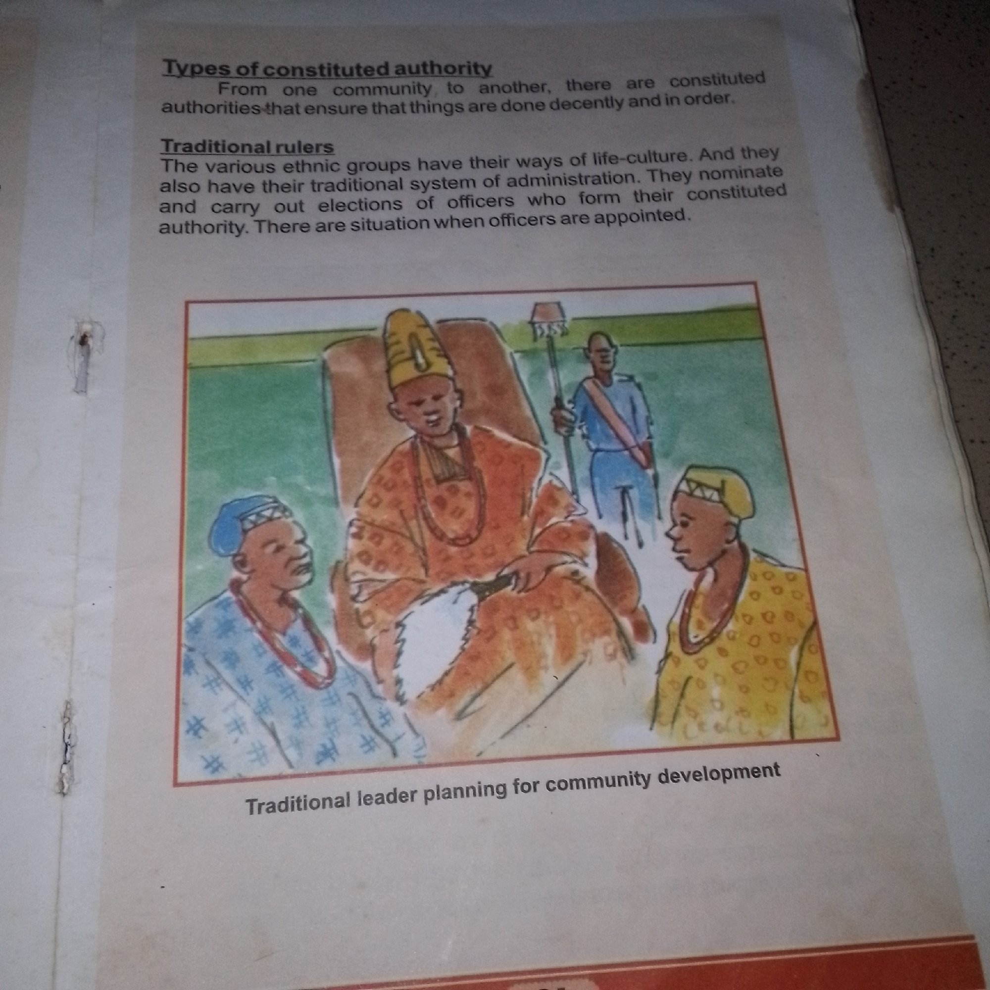 This is a close-up, top-down shot of a page from a book. The page contains text and an illustration. The text sections are titled "Types of constituted authority" and "Traditional rulers." The illustration depicts a scene with several individuals in traditional attire. In the center, a person adorned in an orange robe and a tall headdress sits on a throne. To the left and right of the seated figure are two individuals facing them, one wearing a blue garment with intricate patterns and a blue cap, and the other wearing a yellow patterned garment and a yellow cap. Behind the seated figure, another individual stands holding a staff. The illustration is a watercolor painting with a distinct style. Below the illustration, there is a caption that reads "Traditional leader planning for community development." The page appears to be part of an educational material. The surrounding environment of the page is a muted, neutral tone, suggesting it is lying on a surface or is part of a larger document.