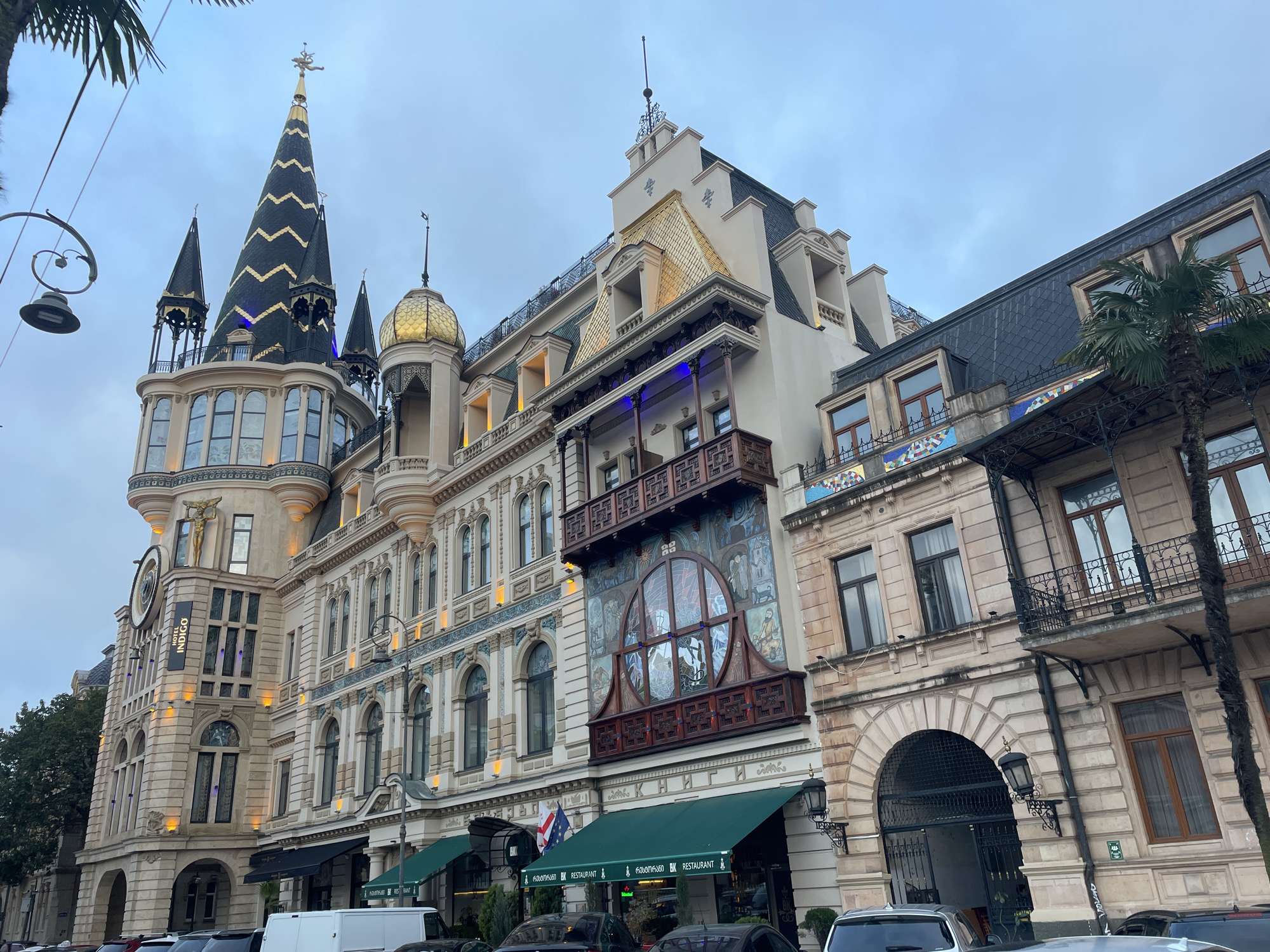 The image depicts a street view in Batumi, Georgia, showcasing a collection of ornate buildings with distinct architectural styles. The building on the left features a tall, slender tower with a dark, patterned roof and gold accents. It is part of the Hotel Indigo, as indicated by signage. Adjacent to it is a building with a lighter facade, intricate window designs, and a balcony with a mural. Next to this building is an older, simpler building constructed of stone.
Various vehicles, including a white van and several cars, are parked along the street in front of the buildings. Green awnings cover the entrances of businesses, including a restaurant. A palm tree and street lamps are visible in the foreground. The sky is overcast and gray.