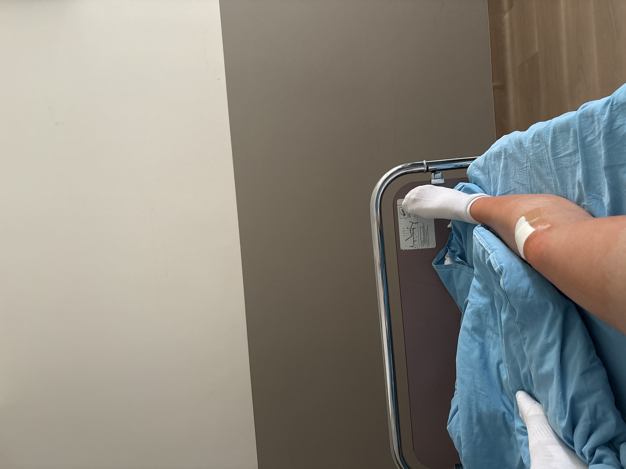 A hospital room is shown, with a bed partially visible on the right side. A person wearing a white sock is lying in the bed, covered by a blue blanket. An IV bandage is visible on the person's arm. The bed's railing is visible on the left. The walls are gray and white, and there are curtains in the background. There is no definitive information to determine if the location is in Sofia, Bulgaria.