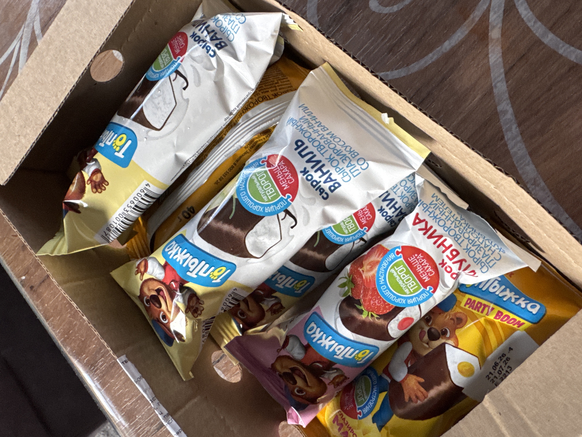 The image shows a close-up view of a cardboard box filled with several individually wrapped ice cream bars. The ice cream bars are of various flavors, including vanilla and strawberry, and feature cartoon bear characters on their packaging. Some of the wrappers have Cyrillic text, including brand names like "Топтыжка" (Toptyzhka) and flavor descriptions such as "СЫРОК ТВОРОЖНЫЙ ГЛАЗИРОВАННЫЙ СО ВКУСОМ ВАНИЛИ" (Curd snack glazed with vanilla flavor) and "СЫРОК ТВОРОЖНЫЙ ГЛАЗИРОВАННЫЙ С НАПОЛНИТЕЛЕМ «КЛУБНИКА»" (Curd snack glazed with strawberry filling). There are also mentions of "PARTY BOOM" and "МЕНЬШЕ САХАРА" (less sugar). The lighting suggests it might be daytime, with shadows indicating sunlight. The setting appears to be indoors, given the cardboard box. No people are visible. The overall impression is a collection of sweet treats, possibly for a party or personal consumption.