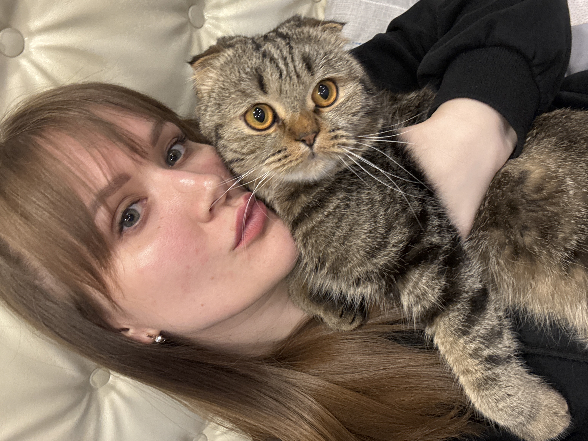 A young woman with long, light brown hair and bangs is captured in a close-up, eye-level shot, lying down with her Scottish Fold cat. The scene takes place indoors, likely in Chaikovsky, Russia, against a backdrop of a light-colored, tufted headboard or cushion.
The woman is positioned on the left, displaying fair skin, light-colored eyes, and a calm expression with slightly parted lips. She wears a black long-sleeved garment, possibly a hoodie, and a small stud earring is visible in her left ear. Her arm is gently around the cat, holding it close to her.
The Scottish Fold cat, resting on the woman's chest and arm, is a prominent subject on the right. It features distinctive tabby markings with shades of brown, grey, and black stripes, and its characteristic folded ears are visible. The cat's large, round eyes are a striking amber-orange, wide open and fixed on the viewer with an alert, perhaps surprised, gaze. Long white whiskers fan out from its muzzle.
The soft, even lighting suggests an indoor environment. The image beautifully conveys a close and affectionate bond between the woman and her feline companion. No text is visible in the media.