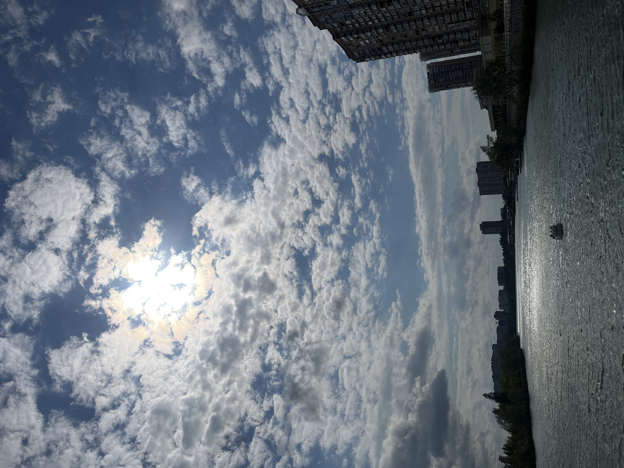 The image shows a body of water reflecting sunlight, with buildings visible along the shoreline. The sky is filled with clouds. Based on the architecture and environment, this is likely in Krasnodar, Russia. A boat is visible in the water.