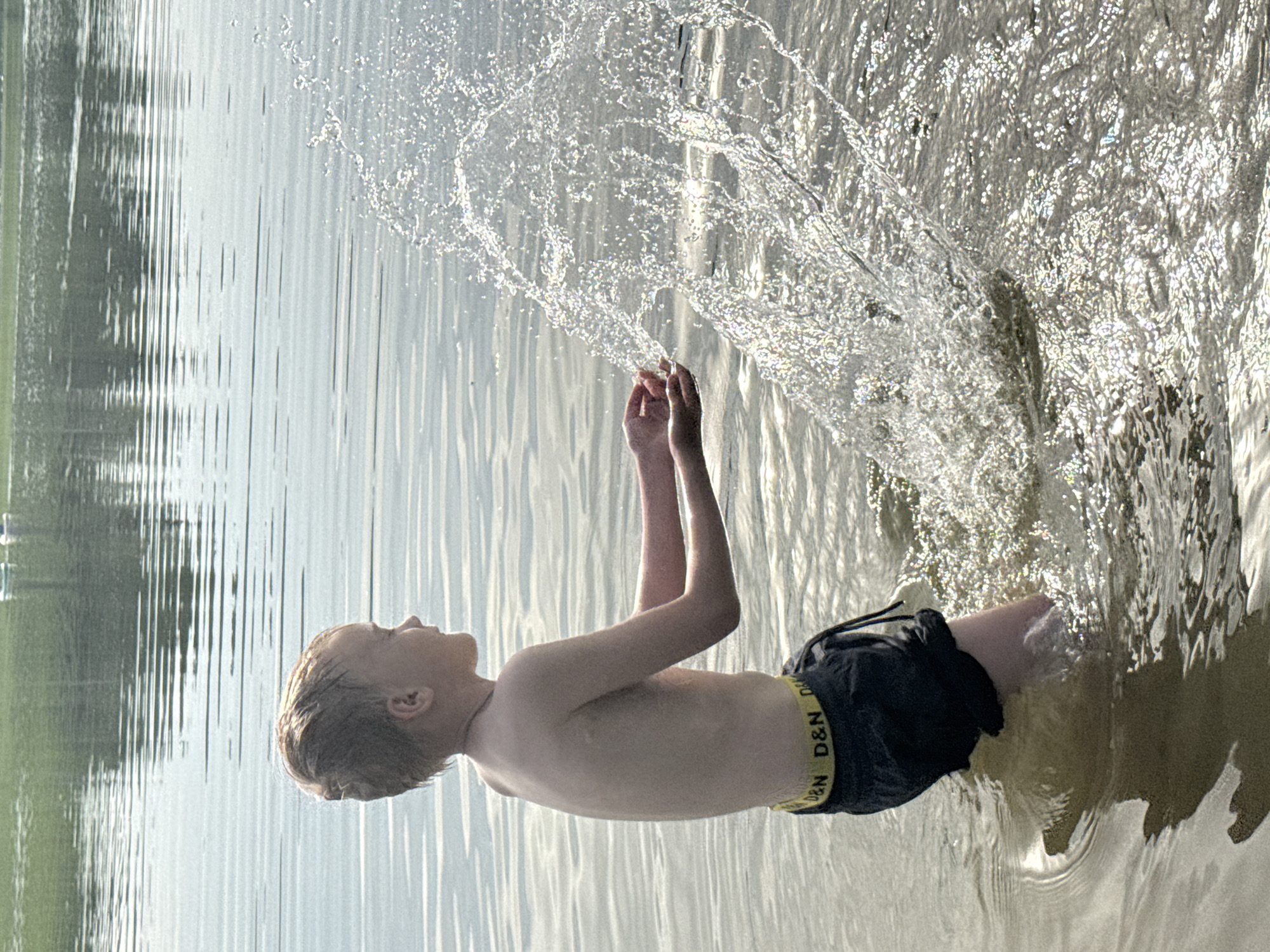 A fair-skinned, blonde-haired boy, wearing dark swim trunks with a yellow band imprinted with "D&N D&N," stands in a body of water, possibly a lake. He appears to be near the shore, based on the change in water color at the edge of the frame. The boy's arms are raised, with water spraying in an arc around them. Trees are visible in the background. The city of Novosinkovo, Russia, cannot be confirmed as the location from the image alone.