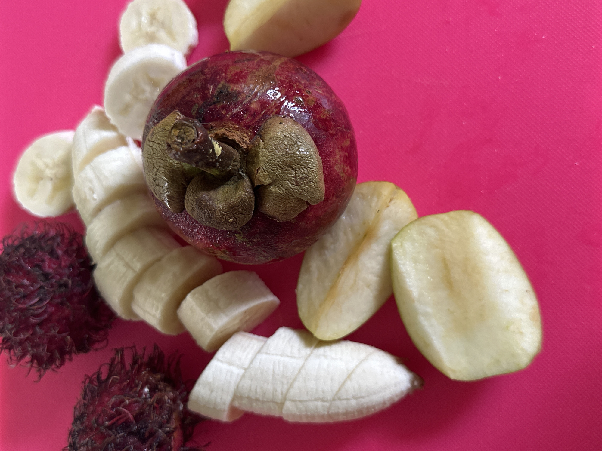 The image depicts a close-up shot of various fruits on a bright pink surface. There is a whole mangosteen with its stem intact, surrounded by slices of banana and what appears to be rambutan fruits. The scene is captured in North Kuta, Indonesia.