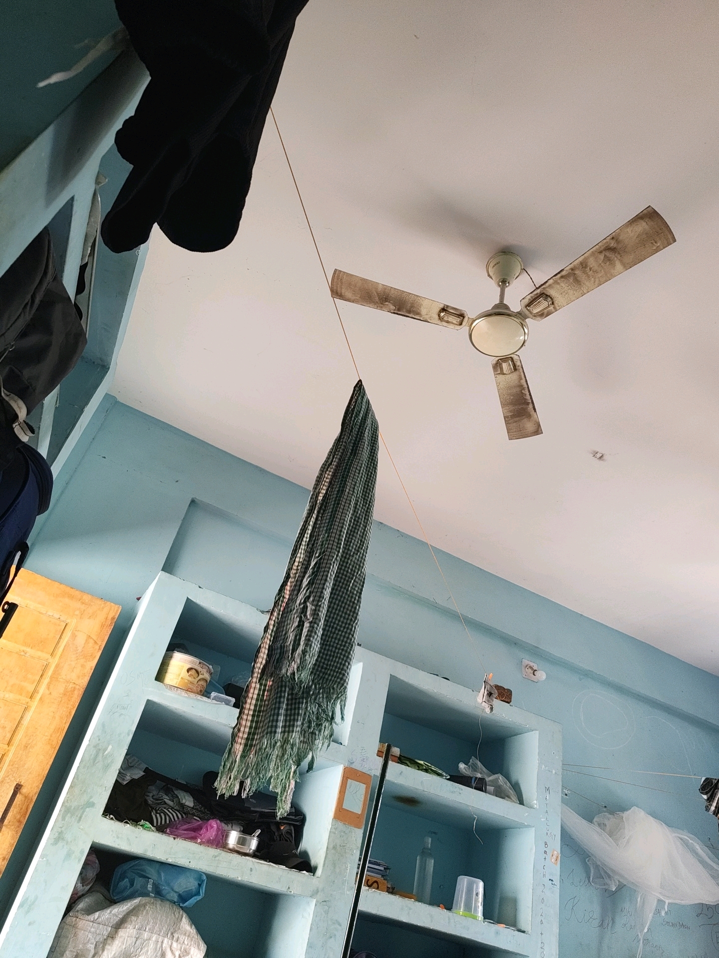 The image depicts an interior scene, likely a room, in Kendujhargarh, India. A ceiling fan, with three dusty blades, is mounted on a white ceiling. A green and white checkered fabric, possibly a scarf or shawl, is hanging from a string that extends from near the fan towards the left side of the frame. In the background, there are light blue shelves built into the wall. The shelves contain various items, including what appear to be bags, containers, and possibly clothing. To the left of the shelves is a wooden door. The walls are painted light blue. Some writing is visible on the wall to the right. The overall impression is of a lived-in, somewhat cluttered space.