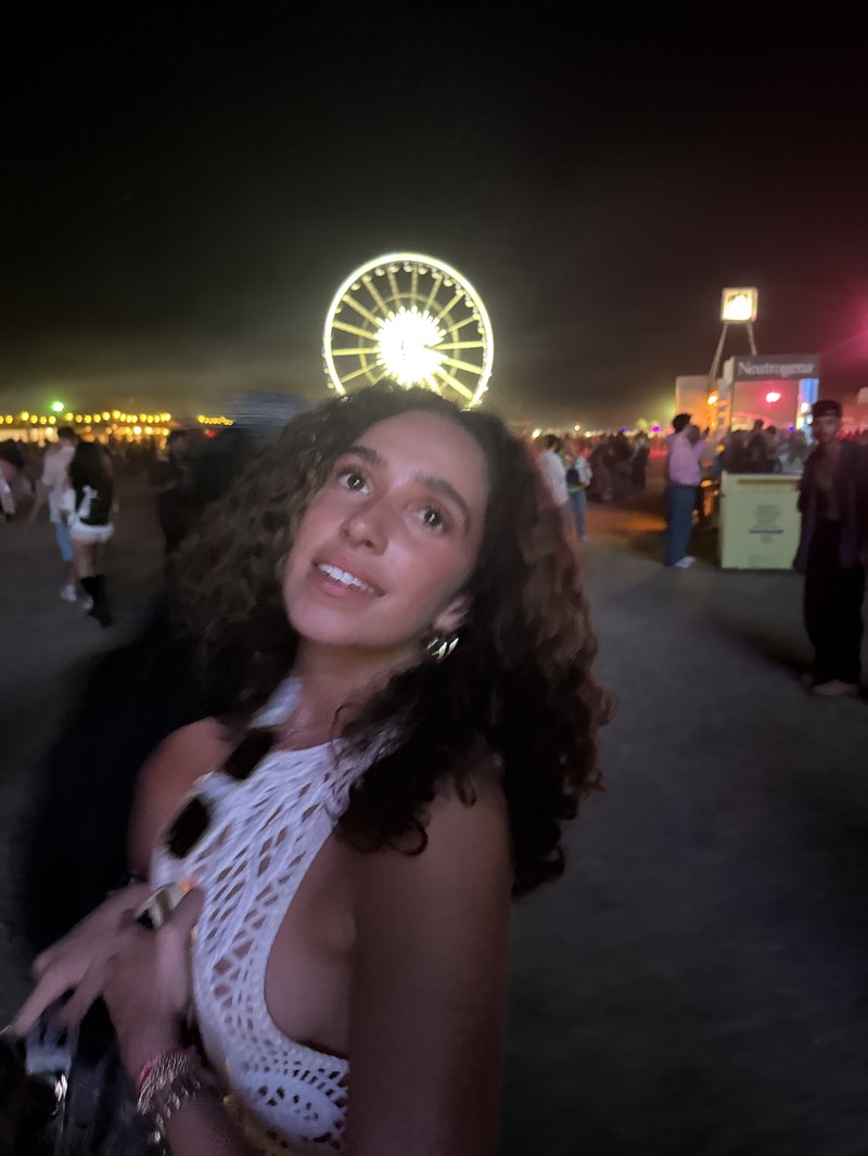 A woman is posing for a picture in Indio, United States, at night. She has curly brown hair and is wearing a white crochet top. A Ferris wheel is visible in the background. There are other people in the background and a Neutrogena tent.