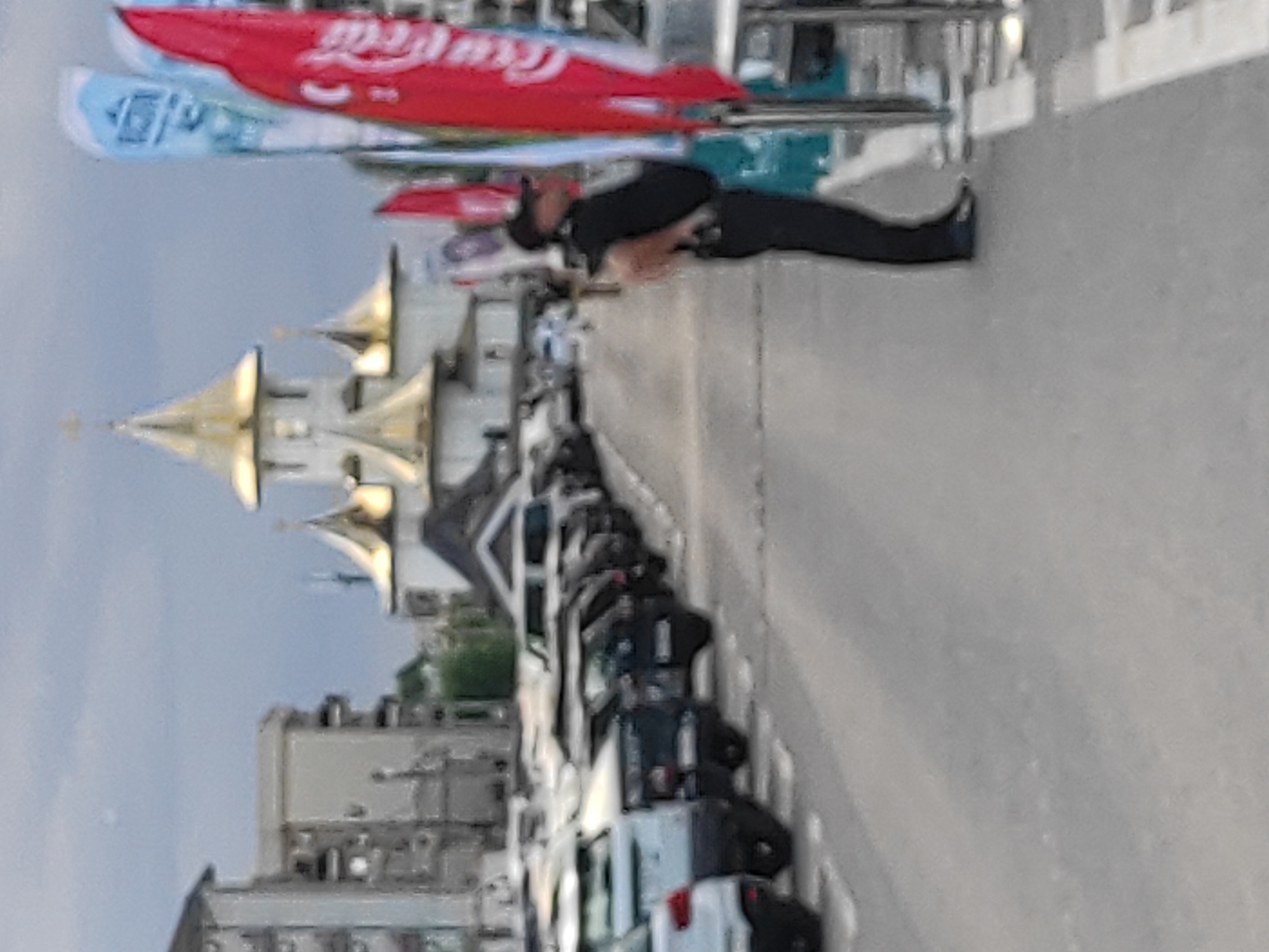 A slightly blurred medium-shot image taken from a low angle shows a parked line of cars in front of a building that appears to be a church with multiple golden domes. A man in a black shirt and black pants, wearing a black baseball cap, stands facing right, with his back to the viewer, near a row of tall flags. One flag, on the right, is red and features the Coca-Cola logo. The scene is set on an asphalt surface, possibly a street or parking lot, with a grey, overcast sky overhead. The background shows apartment buildings. The presence of the church with golden domes and the general atmosphere suggest the location is likely Năvodari, Romania, a coastal city known for its churches.