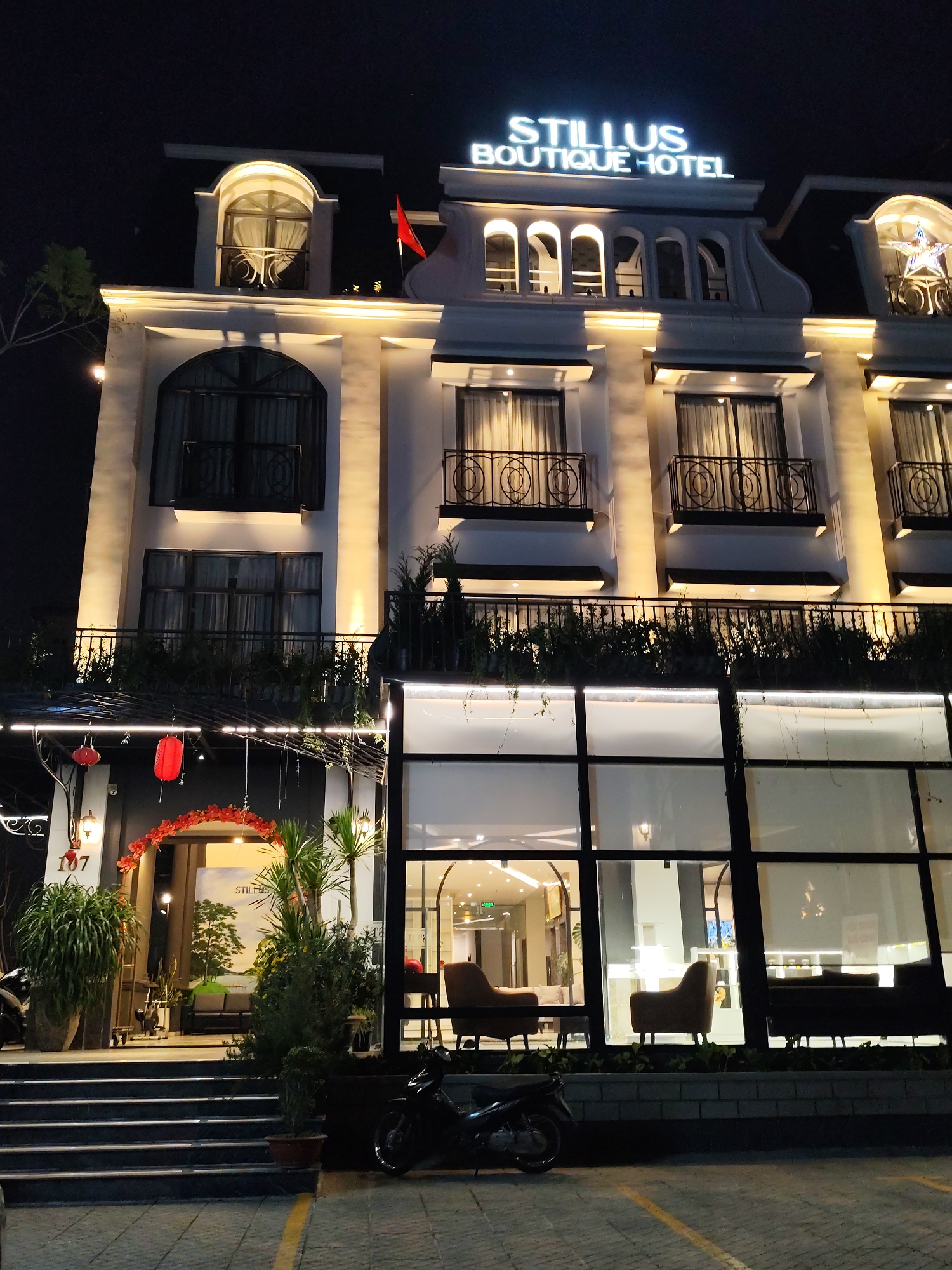 The image shows the exterior of Stillus Boutique Hotel in Da Lat, Vietnam, at night. The hotel is a three-story building with a white facade and decorative balconies with ornate ironwork. The top floor has arched windows, and the roofline is illuminated with the hotel's name in bright lettering. A small red flag is visible on the roof. The lower level has large windows offering a view of the hotel's interior, which includes seating areas. To the left of the main entrance is a decorated archway with red floral arrangements and lanterns. A motorcycle is parked on the paved area in front of the building. The scene is illuminated by the hotel's exterior lighting and ambient light from the street.