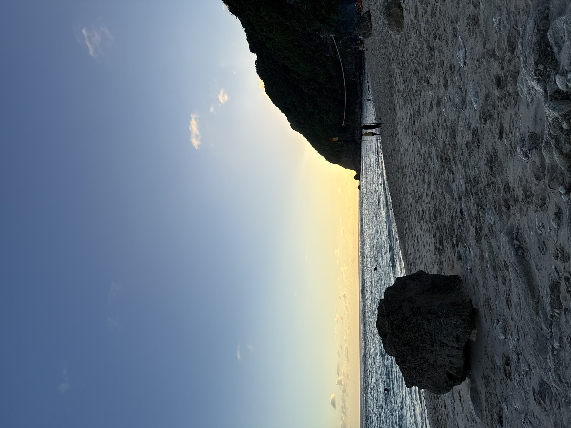 The image depicts a beach scene in Ungasan, Indonesia. A large, dark rock sits on the sandy beach near the water's edge. The ocean stretches along the horizon, reflecting a yellow and orange sunset glow. The sky transitions from a pale blue to a warm yellow. In the distance, a dark green cliff rises along the coast. Faint outlines of people and a volleyball net can be seen further along the beach. The sand is a mix of grey and white. The image is oriented sideways.