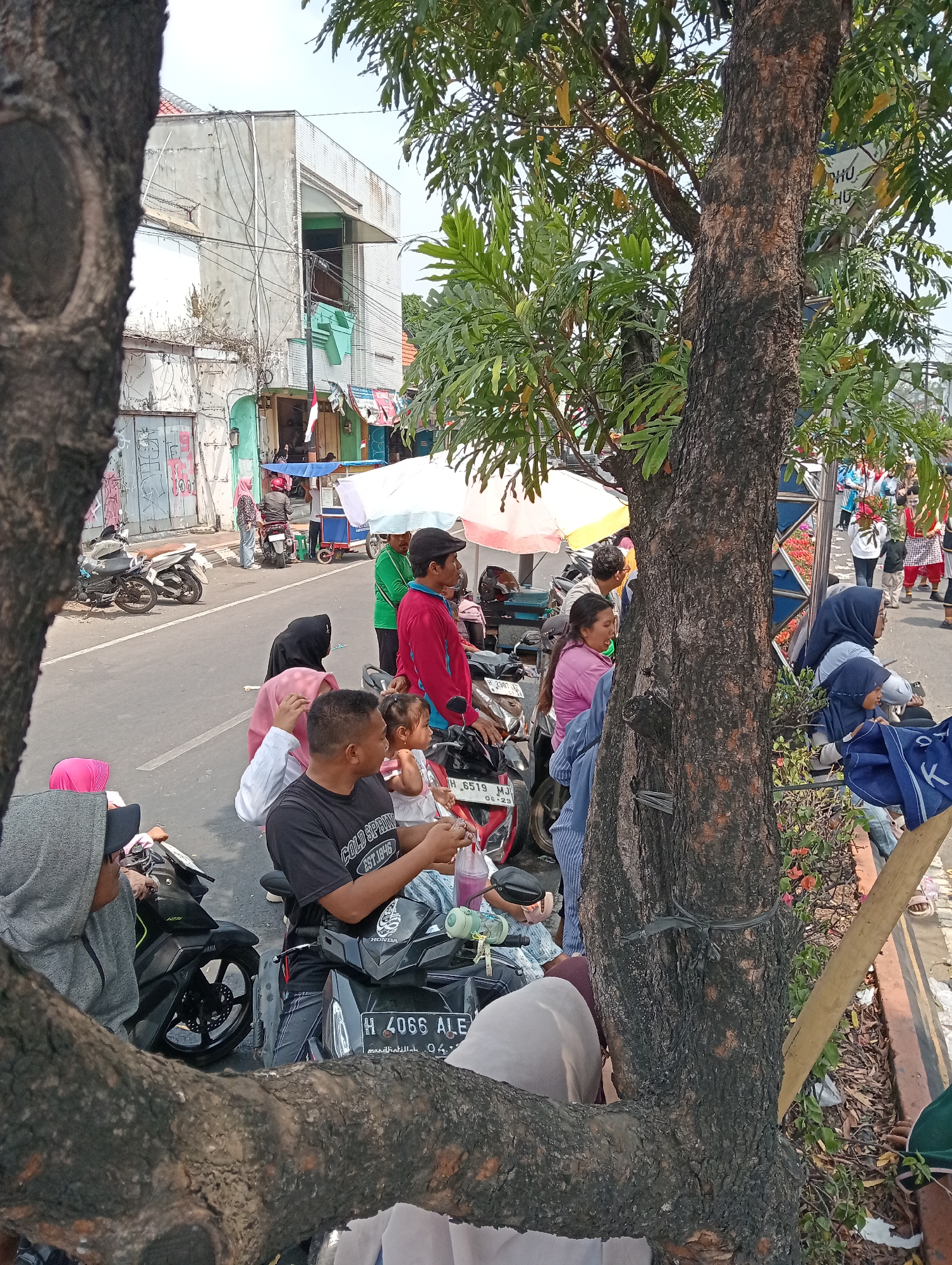 The image depicts a street scene in Demak, Indonesia, with several people gathered around motorcycles. A man wearing a black t-shirt with the text "COLD SPRING EST.1846" sits on a motorcycle, holding a plastic bottle and seemingly interacting with a young child. To his left, a person wearing a grey hooded jacket is seated on a motorcycle. In the background, various individuals are visible, some standing and others on motorcycles. Buildings with visible wiring and a colorful umbrella are present, suggesting a commercial or residential area. The street is paved, and parked motorcycles are a prominent feature of the scene. Trees with thick trunks and green foliage frame the image, casting shadows and adding to the overall atmosphere of a busy outdoor gathering.