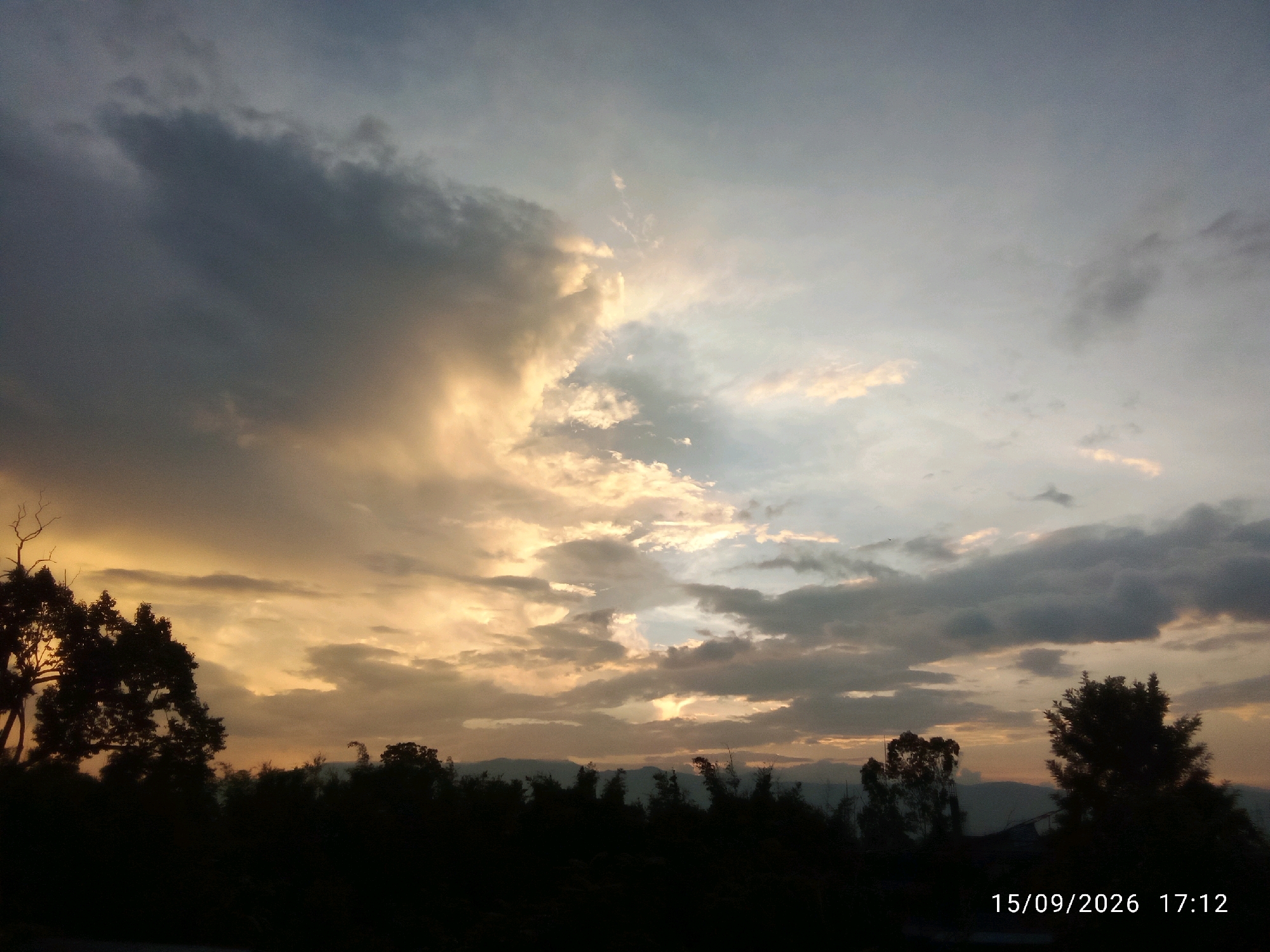 The image depicts a silhouetted landscape under a dramatic sky, likely during sunset or sunrise. Dark trees and foliage form a dense line across the lower portion of the frame, with some taller trees and a single bare branch standing out. In the distance, faint mountain ranges are visible. The sky is filled with a mix of dark, heavy clouds on the left, tinged with warm orange and yellow hues from the setting or rising sun, and lighter, diffused clouds on the right. Shafts of light break through the clouds, illuminating parts of the sky. A timestamp in the bottom right corner reads "15/09/2026 17:12". While the general location is implied to be Patsoi, India, the image itself does not contain specific landmarks or identifiable features that would confirm this. There are no people, animals, or specific structures clearly visible in the scene.