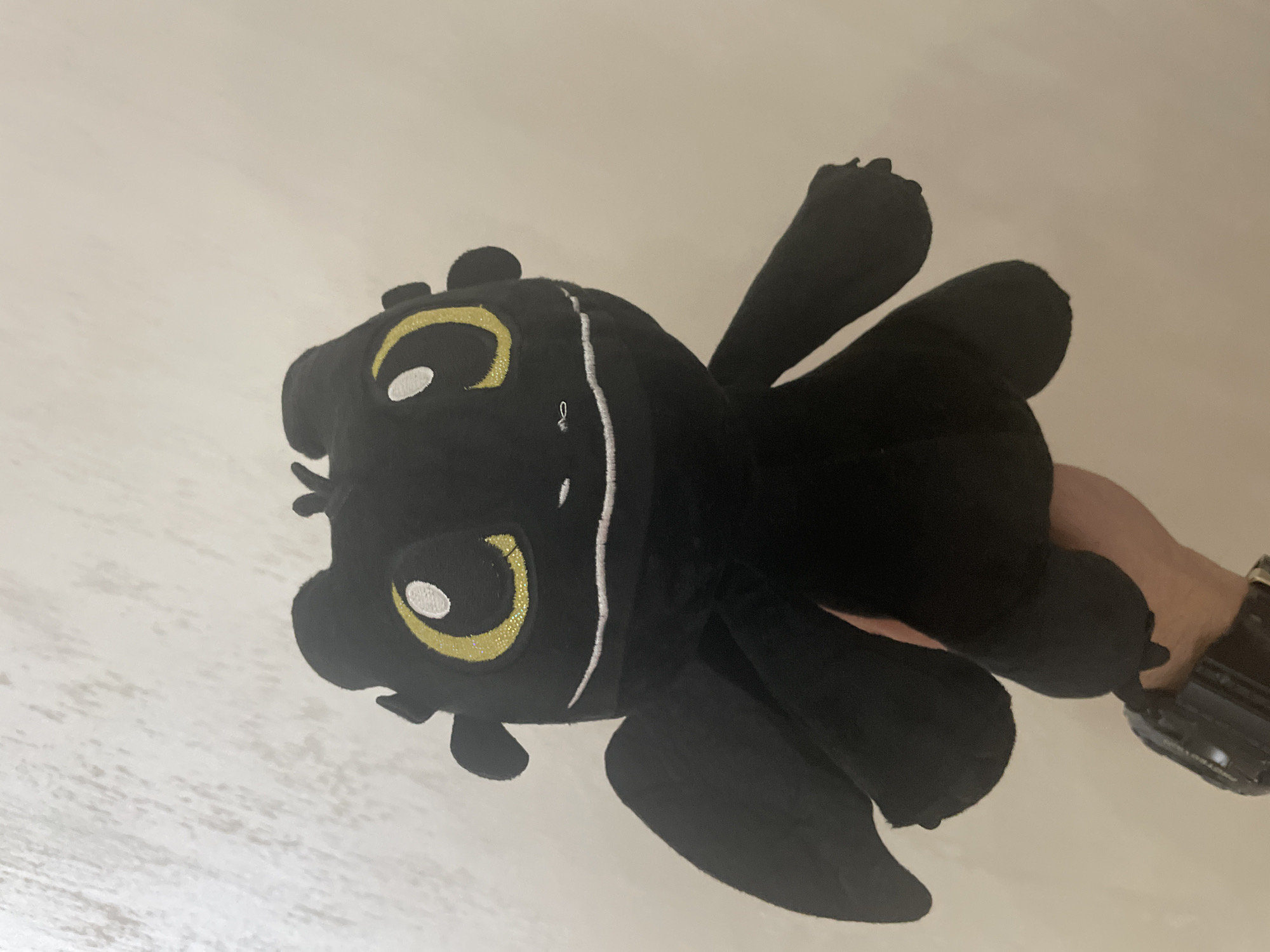 A black plush toy, resembling a dragon with yellow-gold embroidered eyes and a white stitched mouth, is held upright by a human hand against a light, textured wall background in the city of Ihtiman, Bulgaria. The plush toy features small, rounded wings on its back. The hand, visible at the bottom of the frame, is wearing a dark-colored digital watch on its wrist. The background appears to be an off-white or cream-colored wall with faint, irregular vertical and horizontal textures.