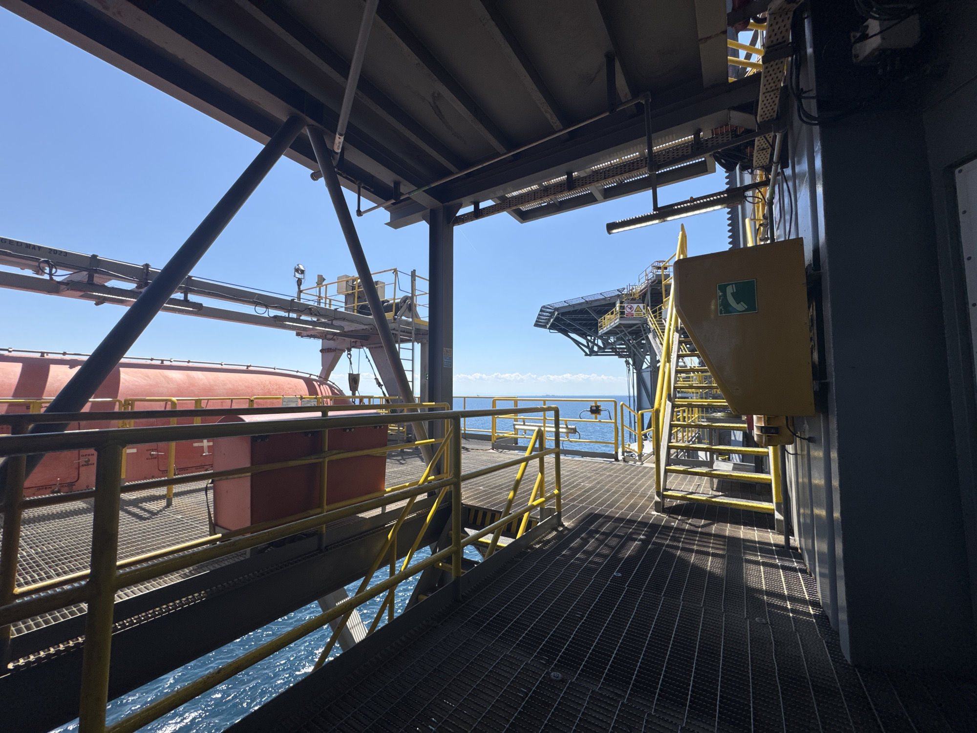 The image shows an outdoor industrial scene on what appears to be an oil rig or similar offshore platform. The perspective is from a walkway with a metal grid floor, looking towards a large red lifeboat suspended from above on the left. Yellow safety railings line the walkway. In the background, the blue ocean is visible under a clear blue sky with scattered white clouds. To the right, there are yellow metal stairs leading up to a higher platform, and a yellow metal enclosure with a green phone symbol on it. The overall environment is maritime and industrial. The location is unknown, Italy.