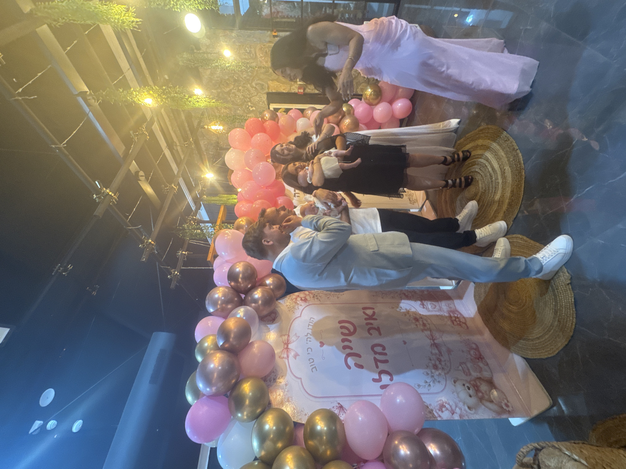 In a dimly lit indoor event space in Migdal HaEmek, Israel, a group of people are gathered around a decorated backdrop. The backdrop, adorned with teddy bears and flowers, features text in Hebrew that appears to be a congratulatory message. Pink and gold balloons arch over the scene. A man in a light blue suit stands to the left of the backdrop, looking down. To his right, a woman in a black dress holds a baby, and another woman in a light-colored dress stands next to her. A woman in a flowing pink dress is on the right side of the frame, facing the group. Several other people are visible in the background, with more balloons and hanging greenery decorating the ceiling. The floor is a dark, reflective surface, possibly marble or tile.