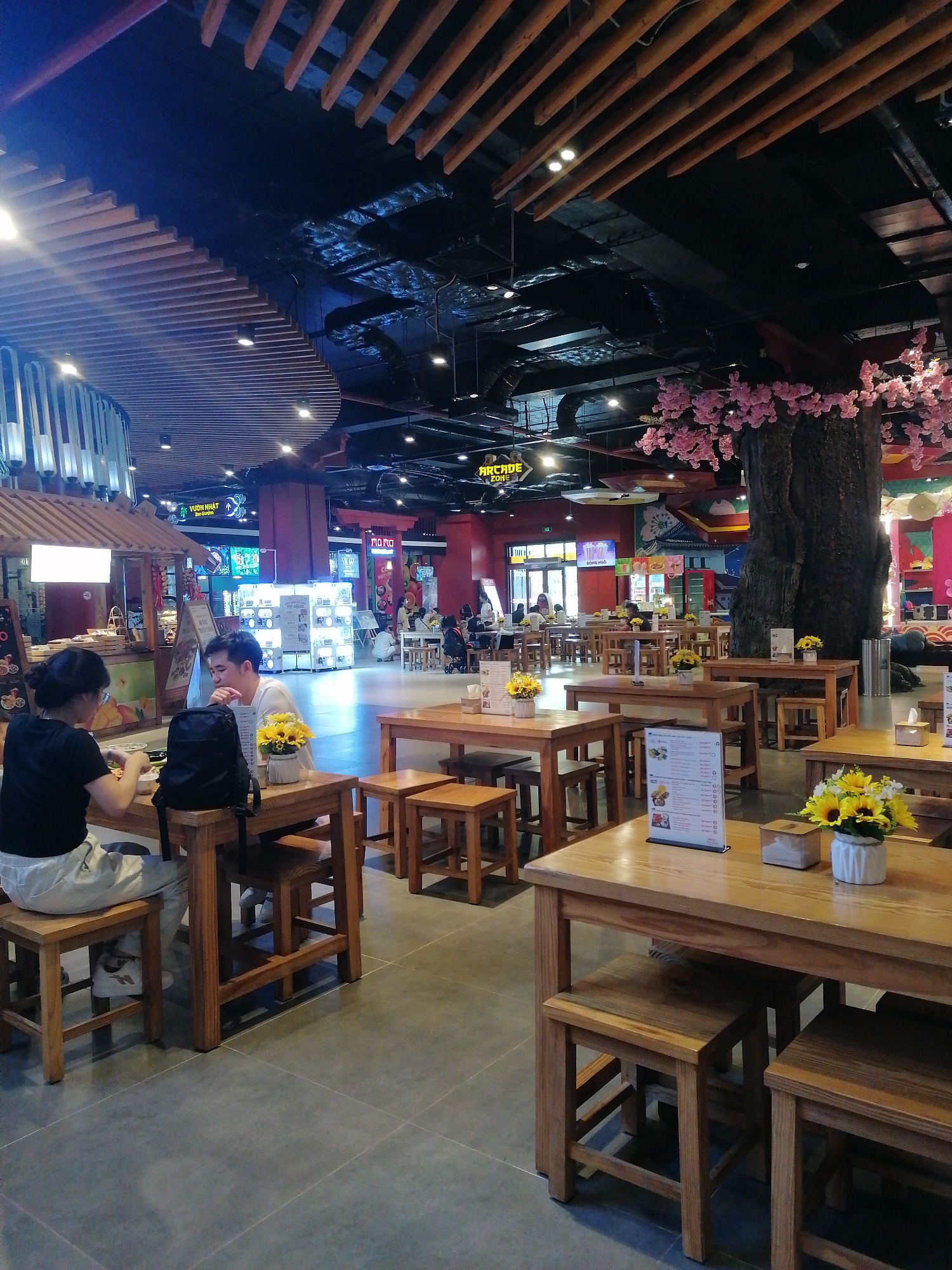 The image shows the interior of a food court in Hong Gai Ward, Vietnam. The space is well-lit with an assortment of wooden tables and chairs arranged for dining. A couple is seated at a table in the foreground, seemingly eating. A black backpack is placed on the table between them. Various other tables are scattered throughout the space, some adorned with floral arrangements in white vases. In the background, there is a large decorative tree with pink flowers. Several storefronts and arcade signs are visible in the distance, indicating the location is part of a shopping center or entertainment complex. The ceiling features a combination of modern lighting fixtures and wooden slat designs.