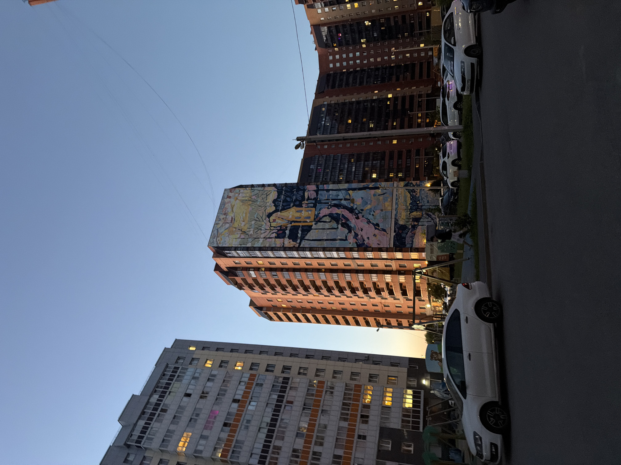 The image depicts a street scene at dusk in front of residential buildings. A white sedan is parked on the left side of the frame. In the background, a tall, red brick apartment building features a large mural on its side depicting a winter landscape with trees and a path. To the left of this building is a grey apartment building with many windows. Several other apartment buildings, some with lights on inside, are visible further in the background. A streetlight and a playground structure with swings are also present. A person is walking on the sidewalk in the middle ground. The sky is a clear, pale blue, indicating the end of the day.