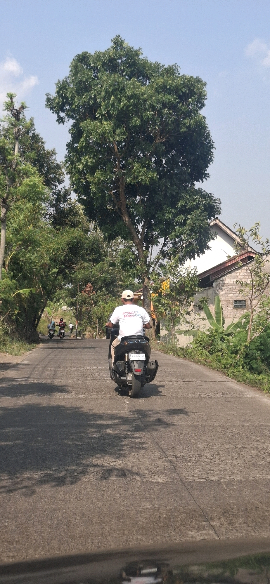 A man wearing a white shirt and a beige cap rides a black scooter down a paved road in Cibinong Hilir, Indonesia. The man's shirt has text that reads "#TONGAY MUNDURAN". The scooter has a license plate that reads "F 4302 W". In the background, two other scooters are visible, as well as a house with a white facade and a brick lower level. Lush green trees line both sides of the road, and the sky is a clear blue with a few white clouds. The road appears to be in a rural or suburban area, with shadows of trees cast across the asphalt.