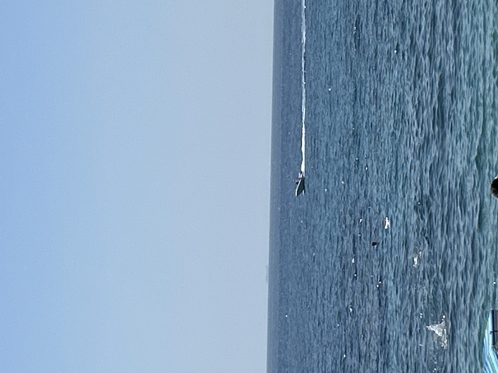 The image depicts a wide expanse of blue ocean under a clear blue sky. In the middle ground, a white speedboat is visible, moving from left to right, leaving a white wake behind it. Further in the distance, on the horizon, a faint, dark shape suggests the presence of land or a larger vessel. Closer to the viewer, in the foreground, the water is a deeper blue with numerous small white reflections or splashes. A hint of a striped beach chair is visible in the bottom left corner, and a dark head is just visible in the bottom center, suggesting people are present at the edge of the water. The overall scene conveys a calm, sunny day at a coastal location.