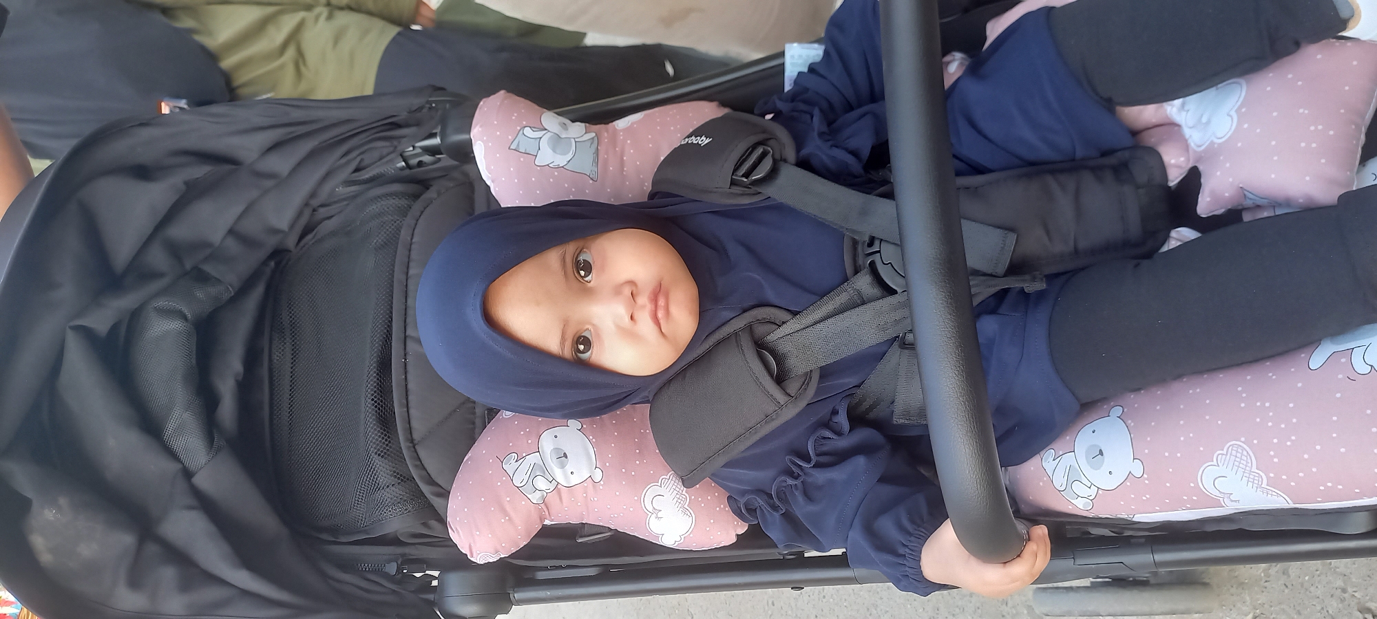 A young child wearing a navy blue hijab and matching long-sleeved top is seated in a black stroller. The child has large, dark eyes and a neutral expression. The stroller has a pink and white patterned liner featuring clouds and cartoon bears, and black safety straps that are buckled. The child's hands are resting on the stroller's handlebar. In the background, two adults are partially visible, suggesting an outdoor public setting. The overall impression is that this is a snapshot of a child in a stroller, likely taken in a public area in Samarinda, Indonesia, given the context provided.