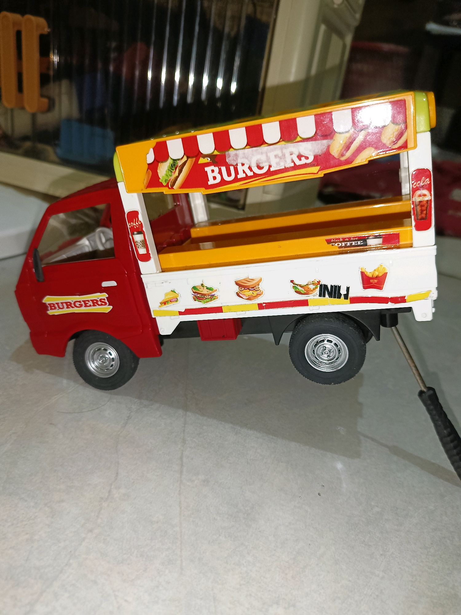 A toy food truck, predominantly red and white, is displayed on a light-colored, tiled floor. The food truck features graphics of burgers, hot dogs, sandwiches, french fries, and drinks, suggesting it is a mobile food vendor. The truck has a yellow interior and a striped awning that reads "BURGERS." The background shows a blurred interior environment with vertical elements, possibly shelves or blinds. A small silver tool with a black handle is visible near the rear wheel of the truck, casting a shadow. There is no indication of the city of Tangerang, Indonesia, within the image itself.