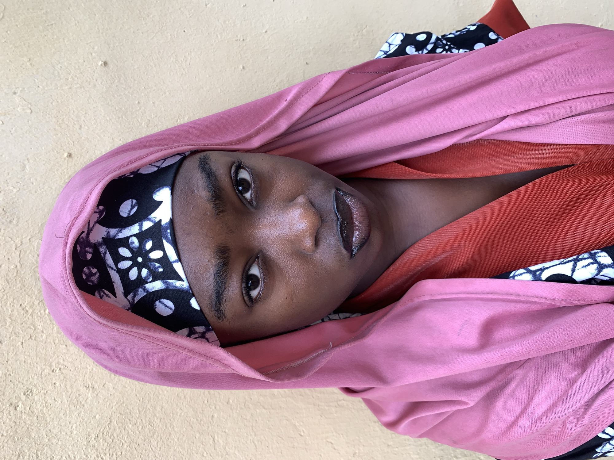 A medium, close-up shot of a woman posing in what looks like an enclosed space with a yellow-tinged wall. She is wearing a pink hijab over an inner layer of black and white patterned fabric. An orange fabric garment appears as a second layer below the hijab. The woman's head is angled towards the top right, and she is looking directly at the viewer.
Because the context does not provide visual elements unique to the city, I cannot provide location information specifically for Rafin Par, Nigeria.