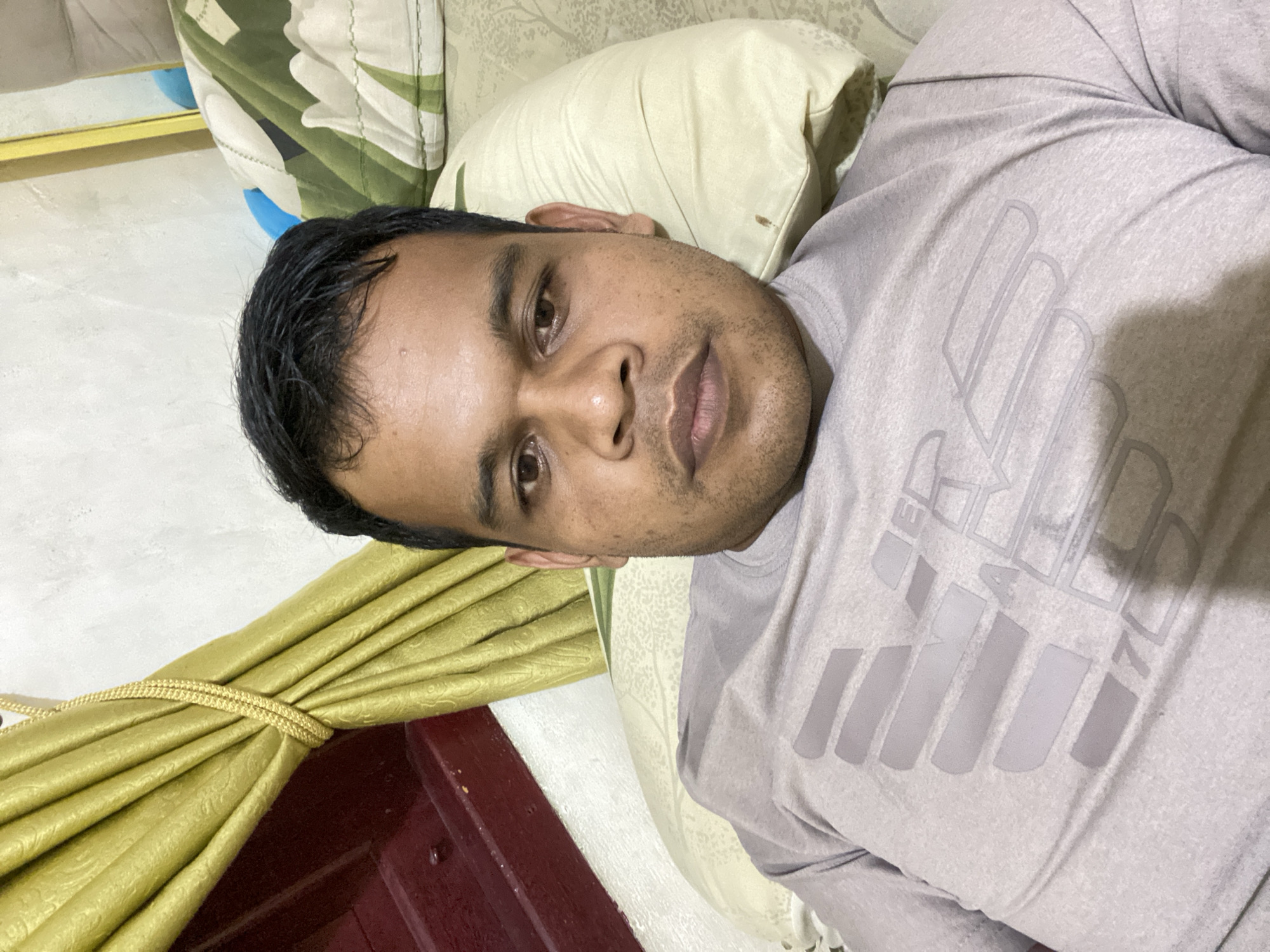 A man is lying down on a bed, taking a selfie. He has short, dark hair and is looking directly at the camera. He is wearing a light purple t-shirt with a grey logo on it. Behind him, there are pillows and a green and white patterned duvet. To his left, there is a draped golden curtain and a dark red wooden surface. The wall behind him is white. The image appears to be taken indoors.